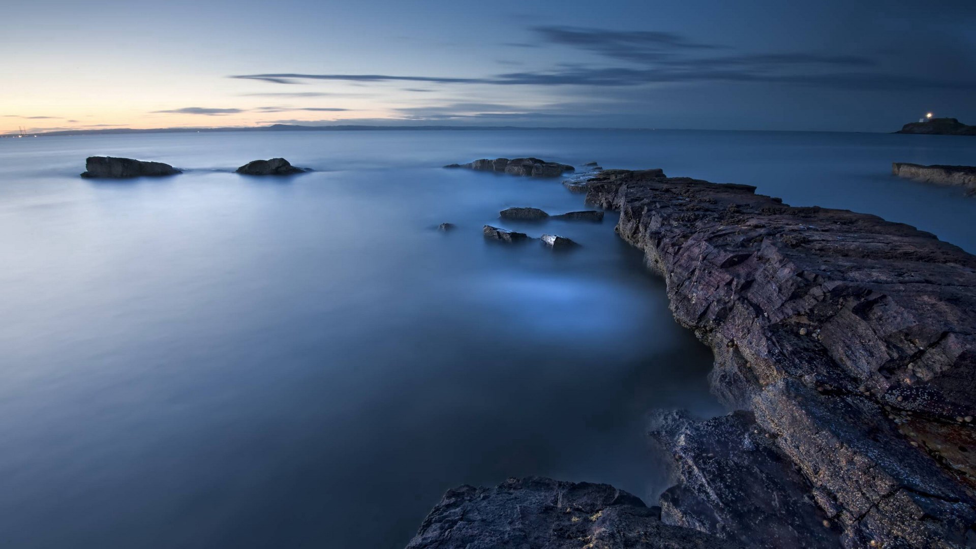 Rocky coastline at dusk with a distant lighthouse HD Wallpaper