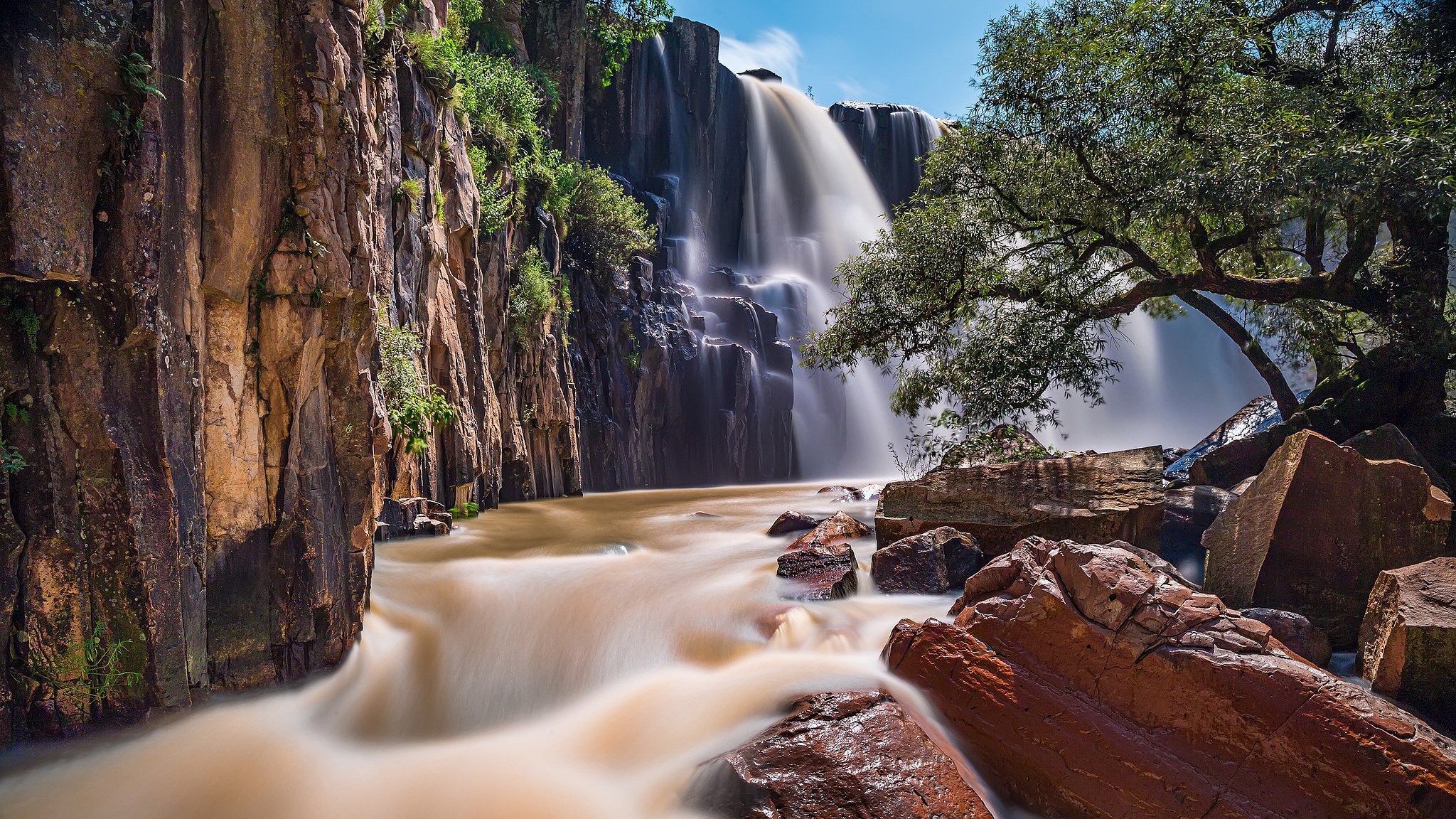 Waterfall cascading over dark rocks with a tree HD Wallpaper