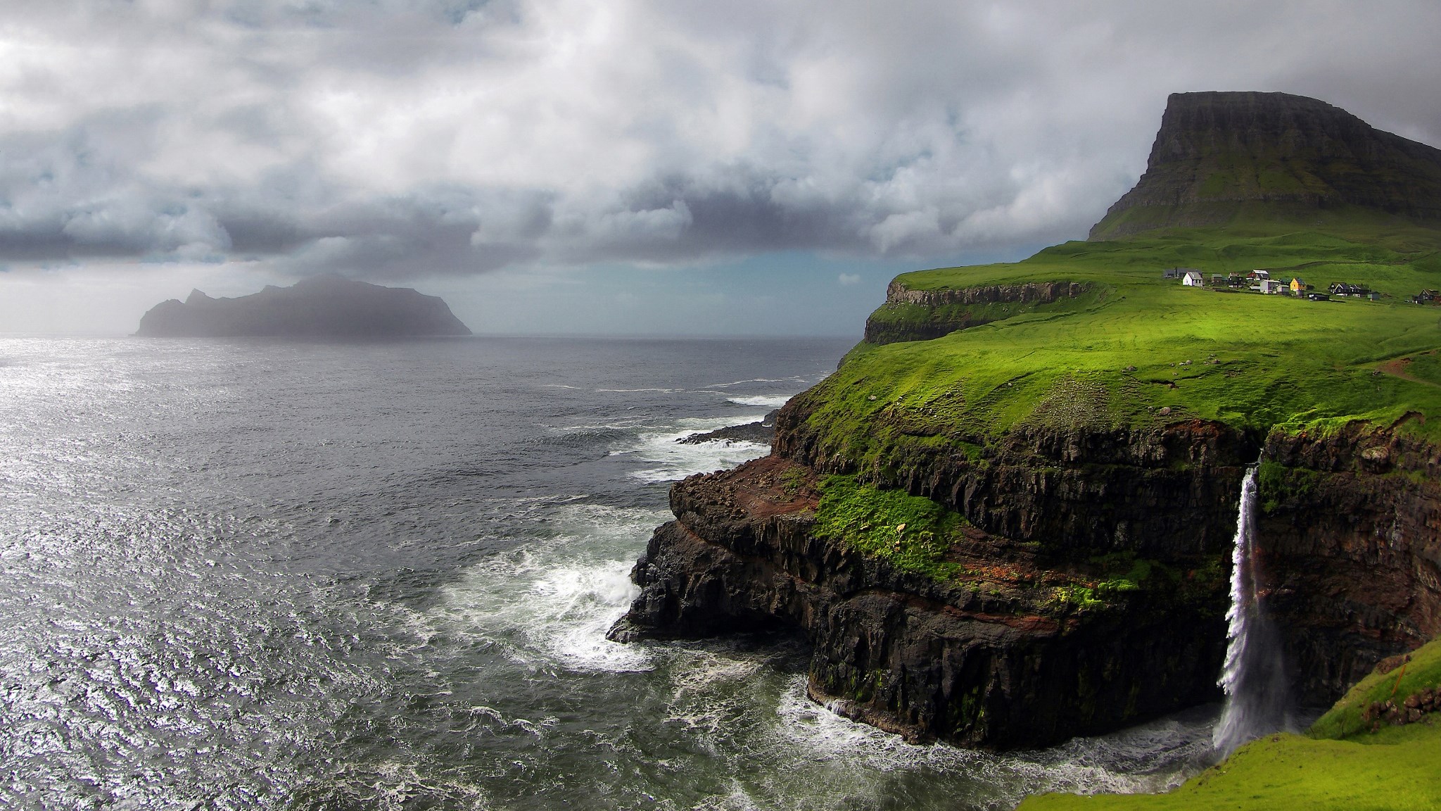 Waterfall cascading from green cliffs into the ocean near a village HD Wallpaper