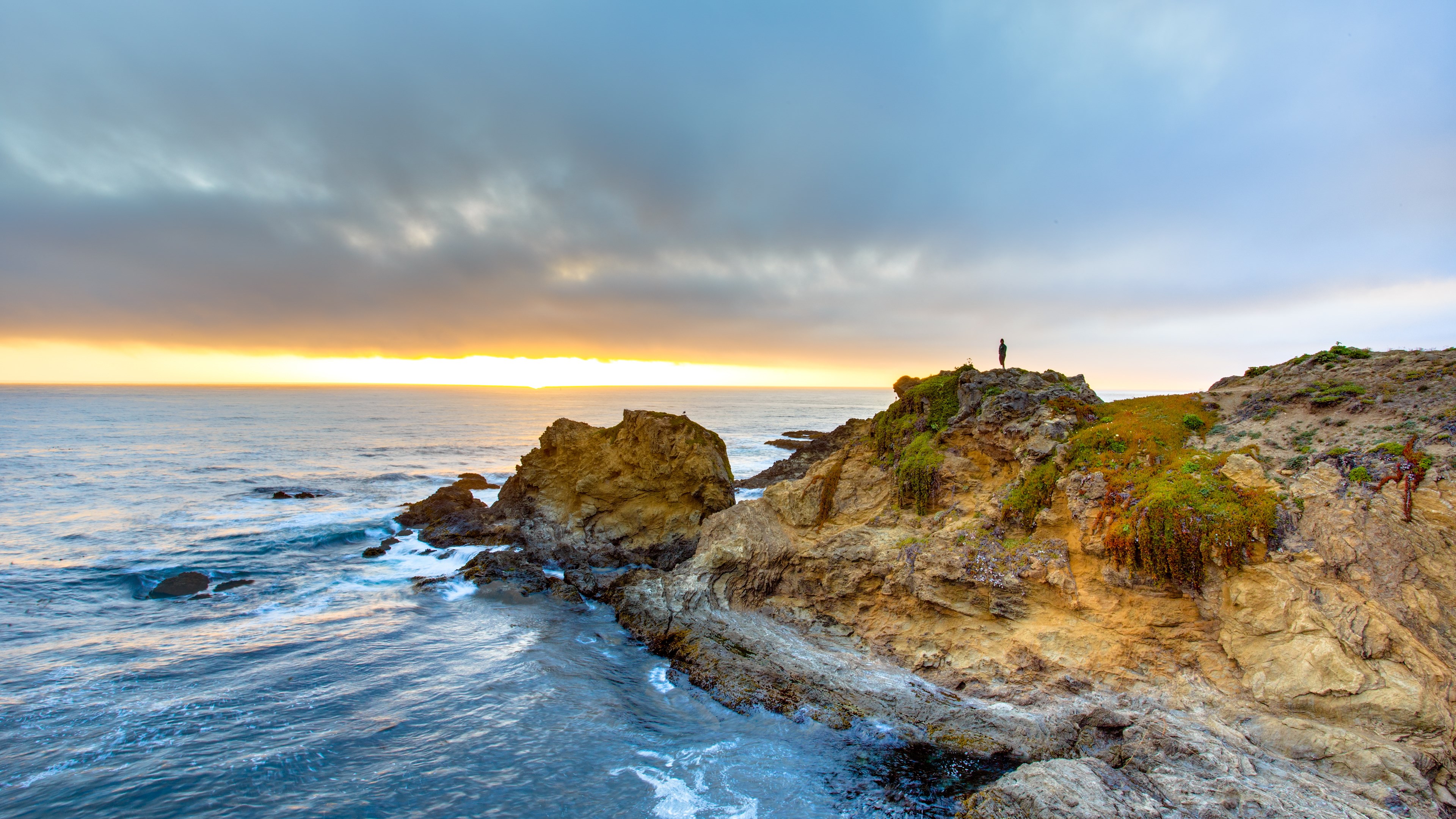 Person standing on a rocky cliff overlooking the ocean at sunset HD Wallpaper