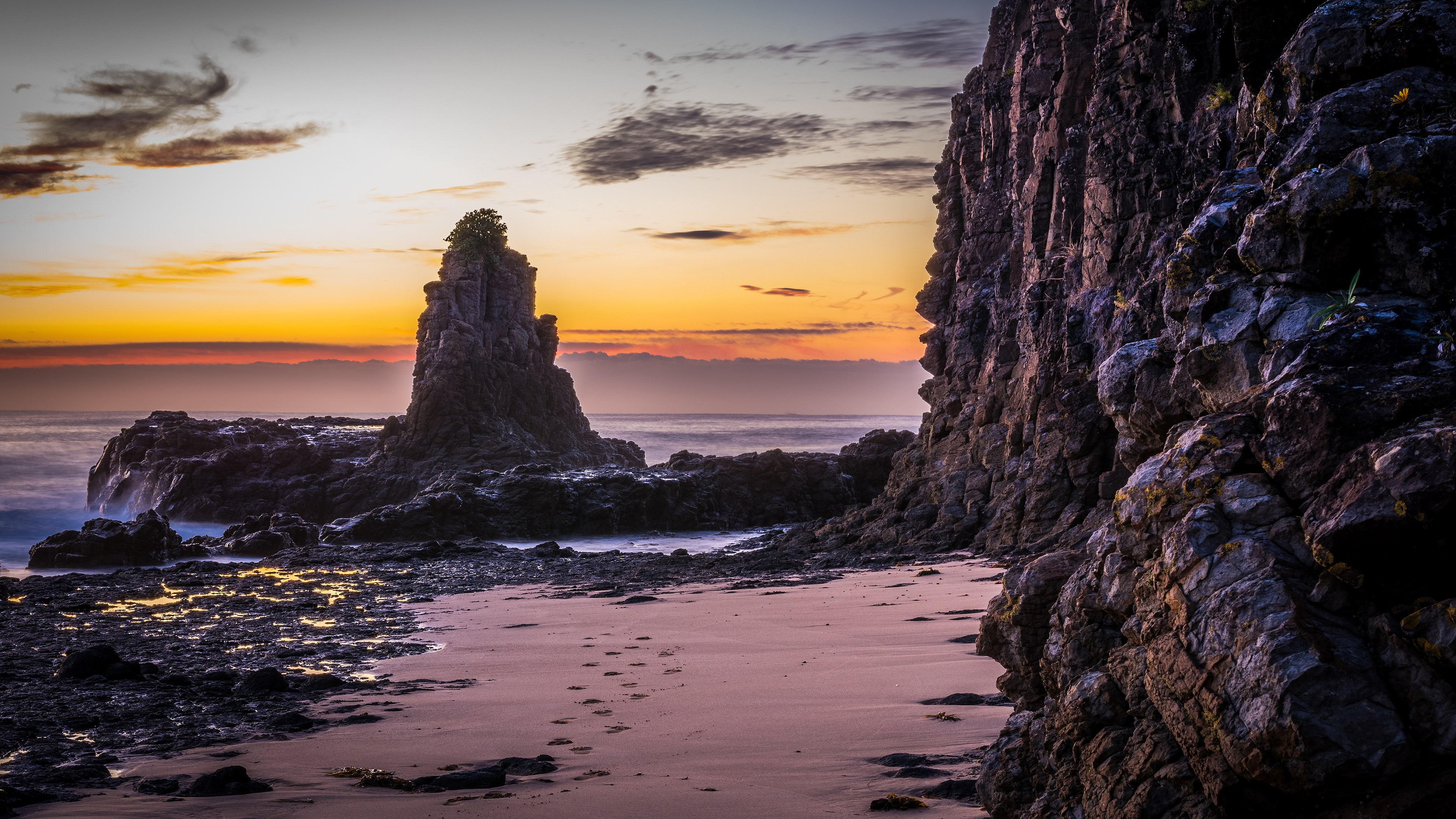 Rocky coastline at sunset with footprints on sand HD Wallpaper