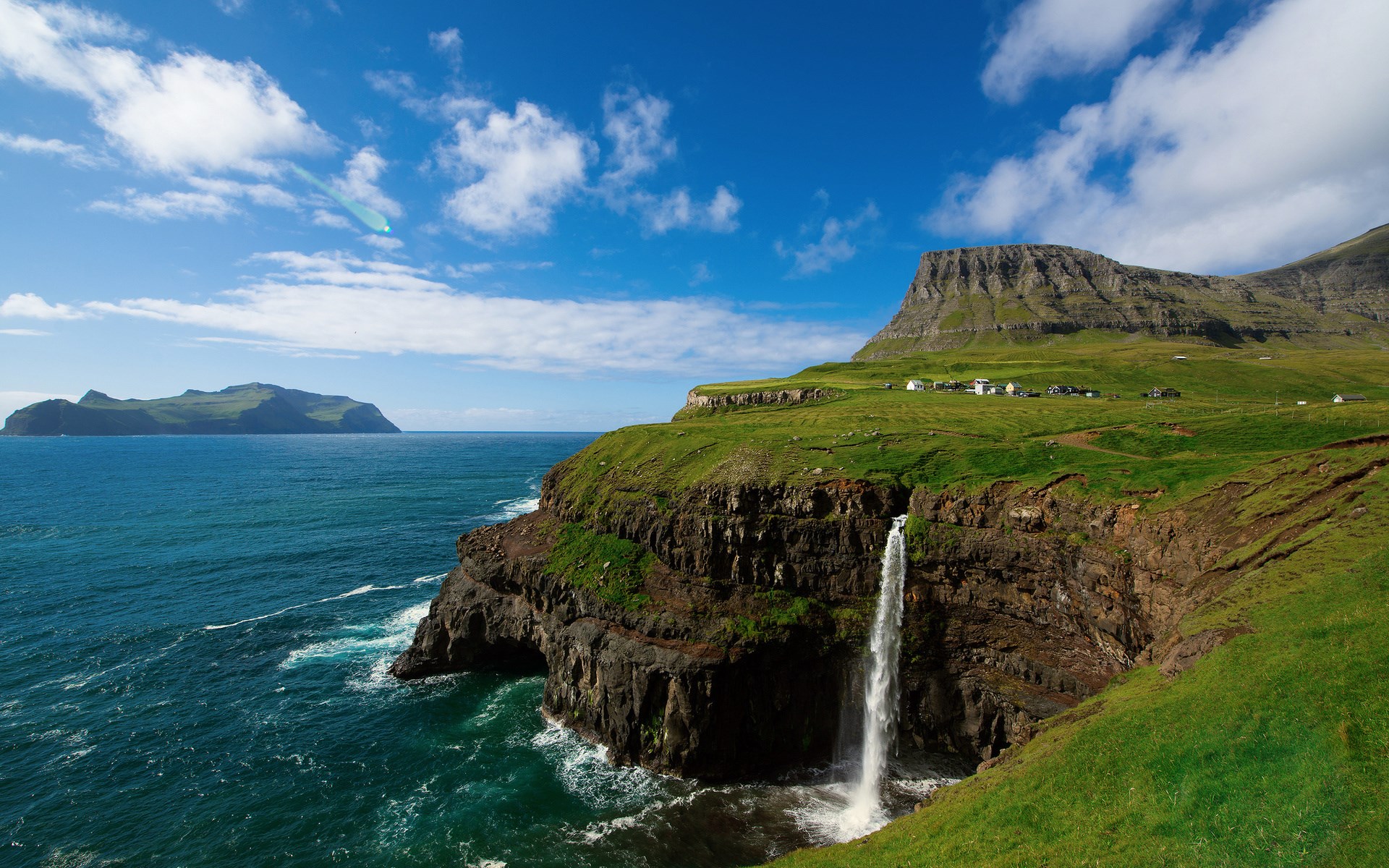 Waterfall cascading from a cliff into the ocean with a distant island HD Wallpaper