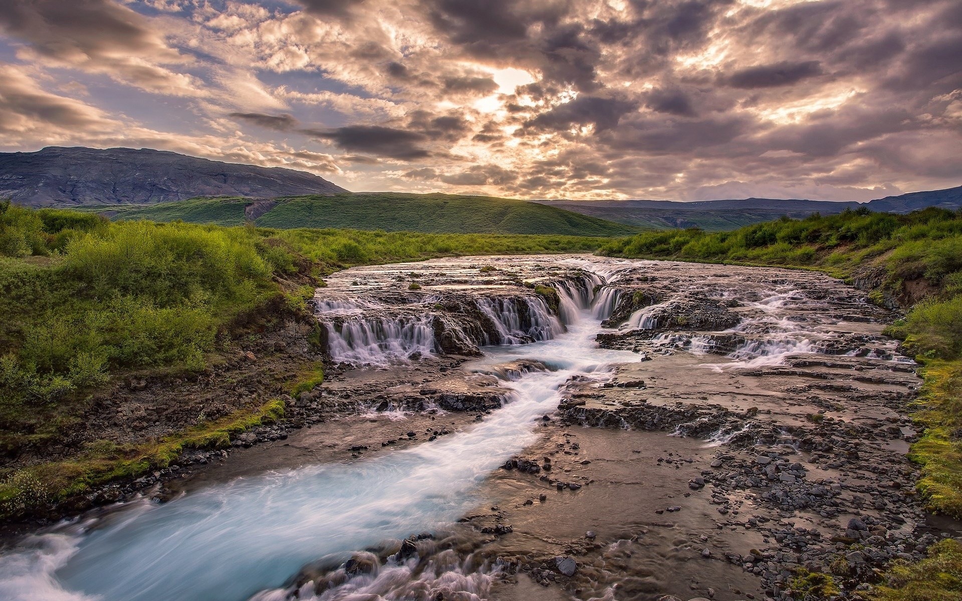 Waterfall flowing over rocks with green hills and cloudy sky HD Wallpaper