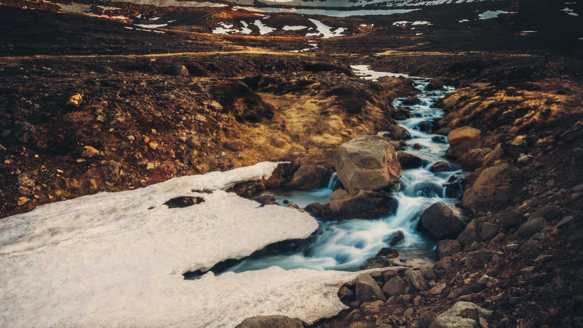Mountain stream flowing through rocky terrain with snow patches HD Wallpaper