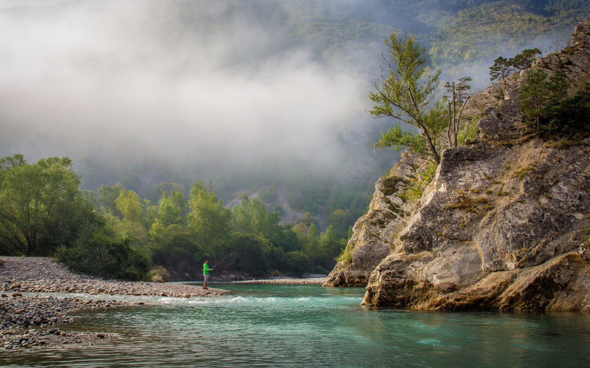 Man fishing in a river with misty mountains HD Wallpaper