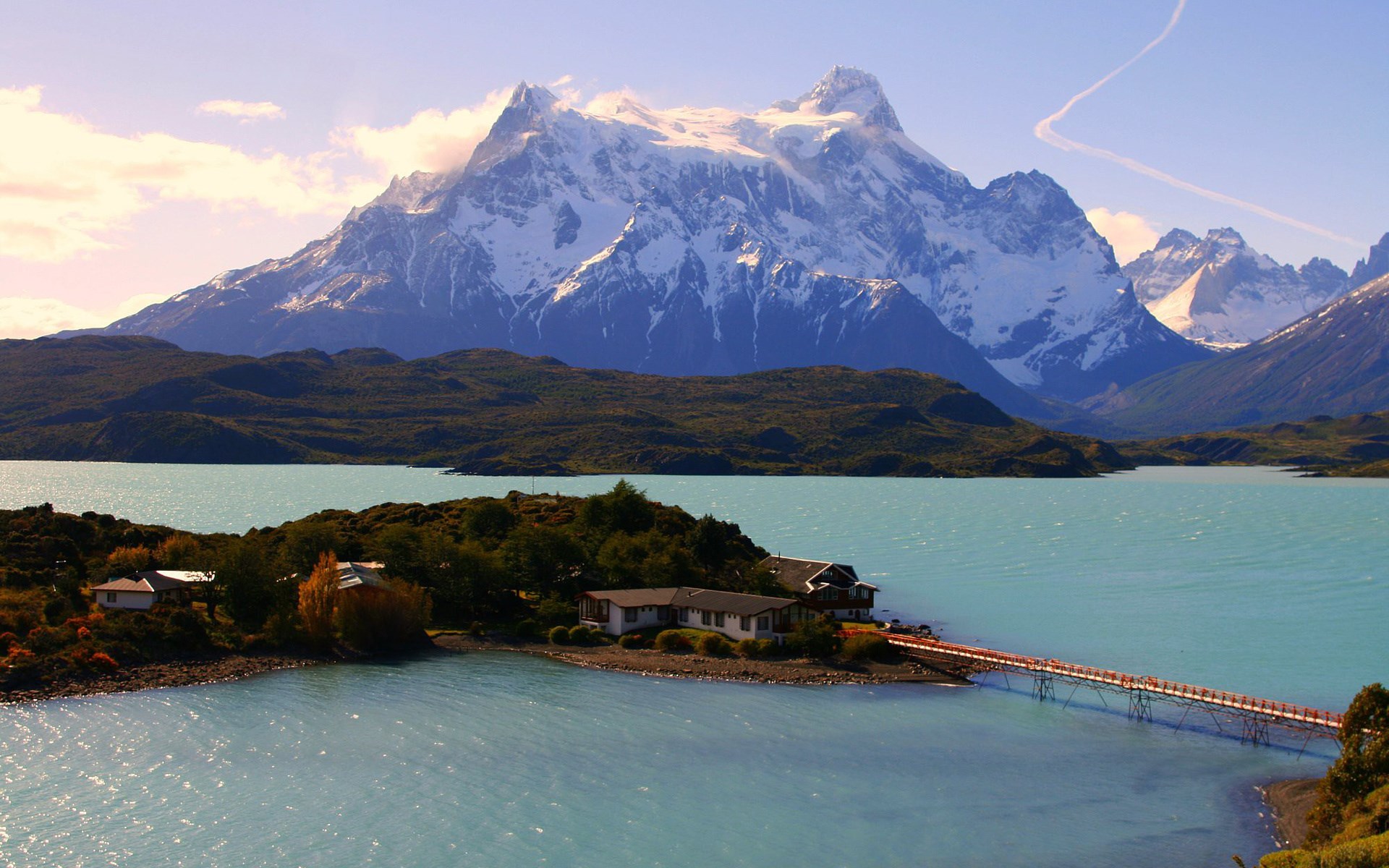 Snow-capped mountains over a turquoise lake with houses and a bridge HD Wallpaper