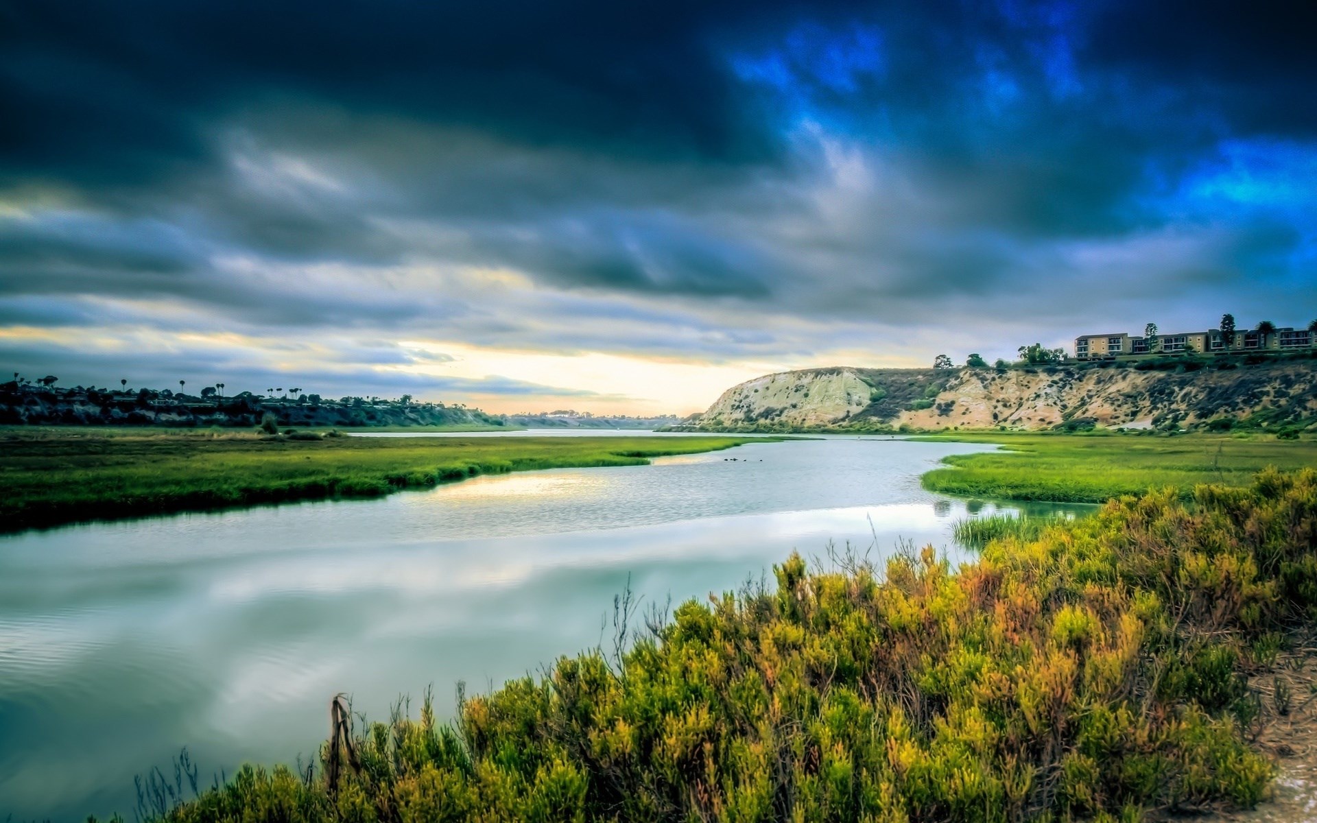 River flowing through marshland with buildings on a hill HD Wallpaper