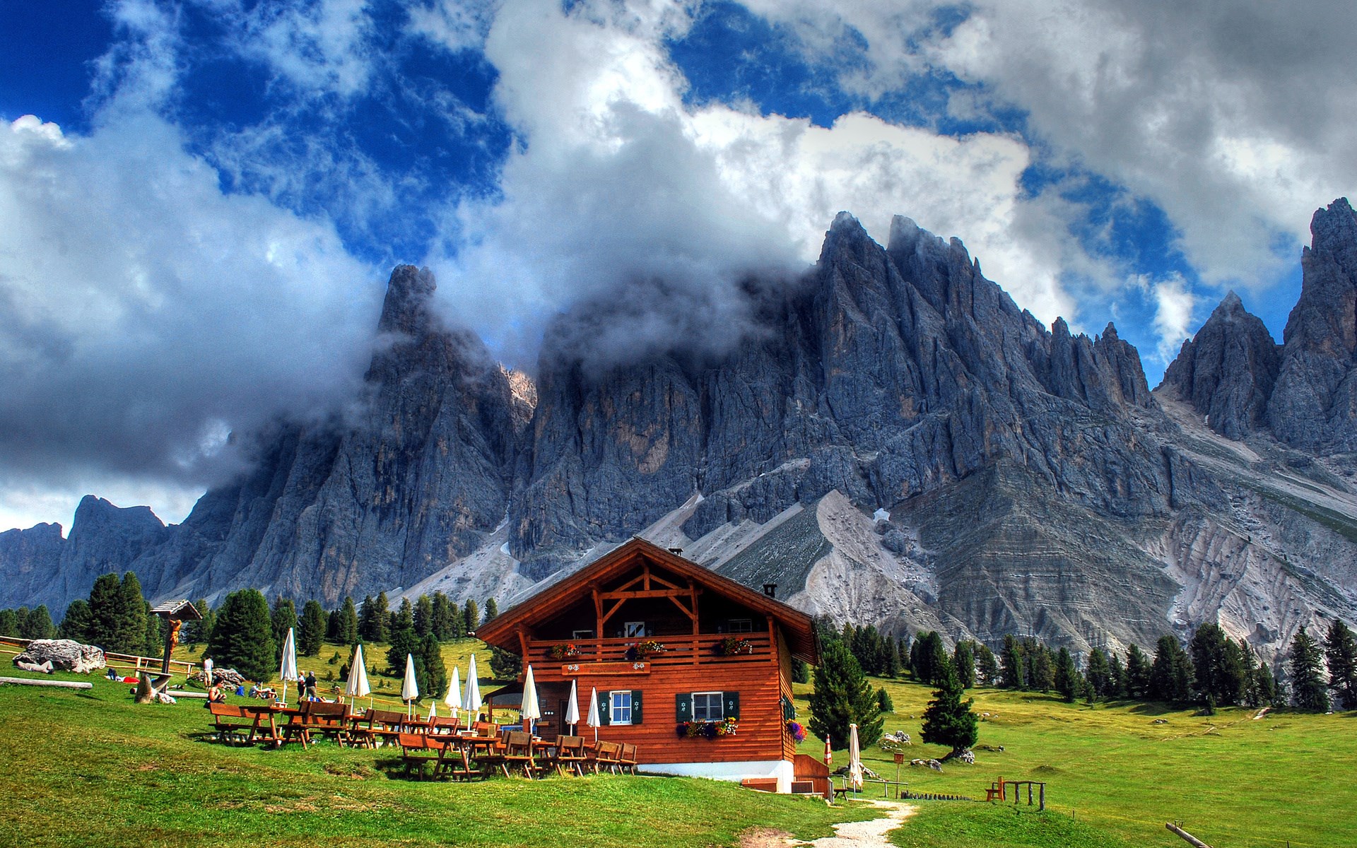 Wooden mountain lodge with outdoor seating and rocky peaks HD Wallpaper