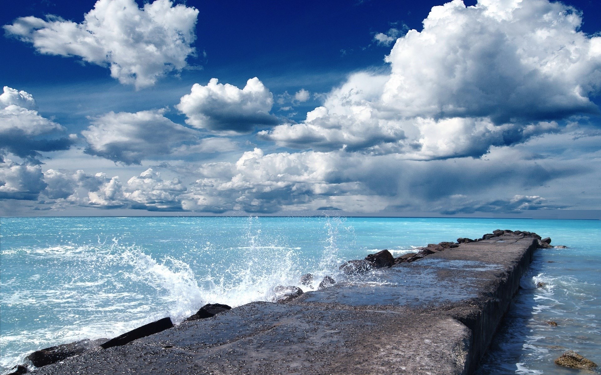 Ocean waves crashing on a concrete pier under a cloudy sky HD Wallpaper