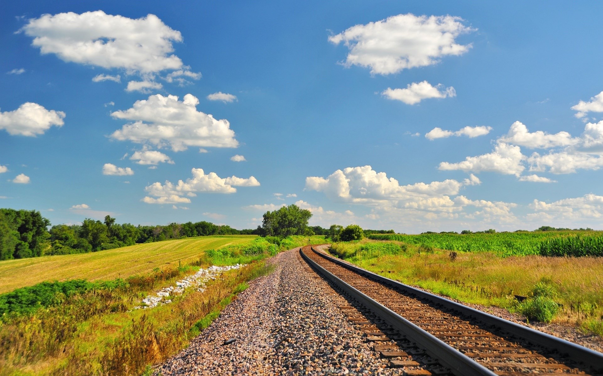 Train tracks curve through a rural landscape with fields and clouds HD Wallpaper