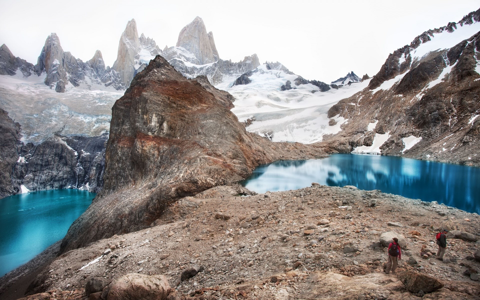 Two hikers near a bright blue glacial lake and jagged mountains HD Wallpaper
