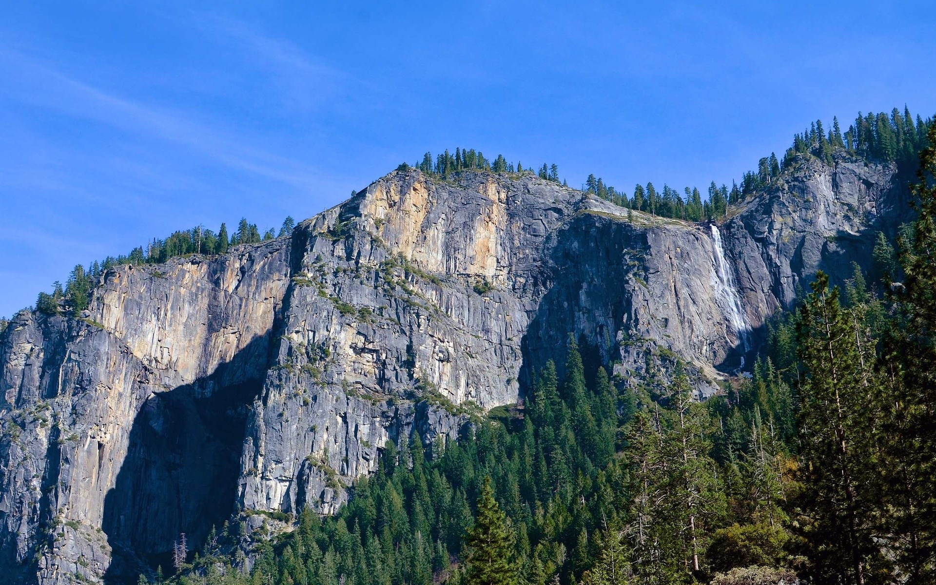 Waterfall cascading down a rocky mountain face with pine trees HD Wallpaper