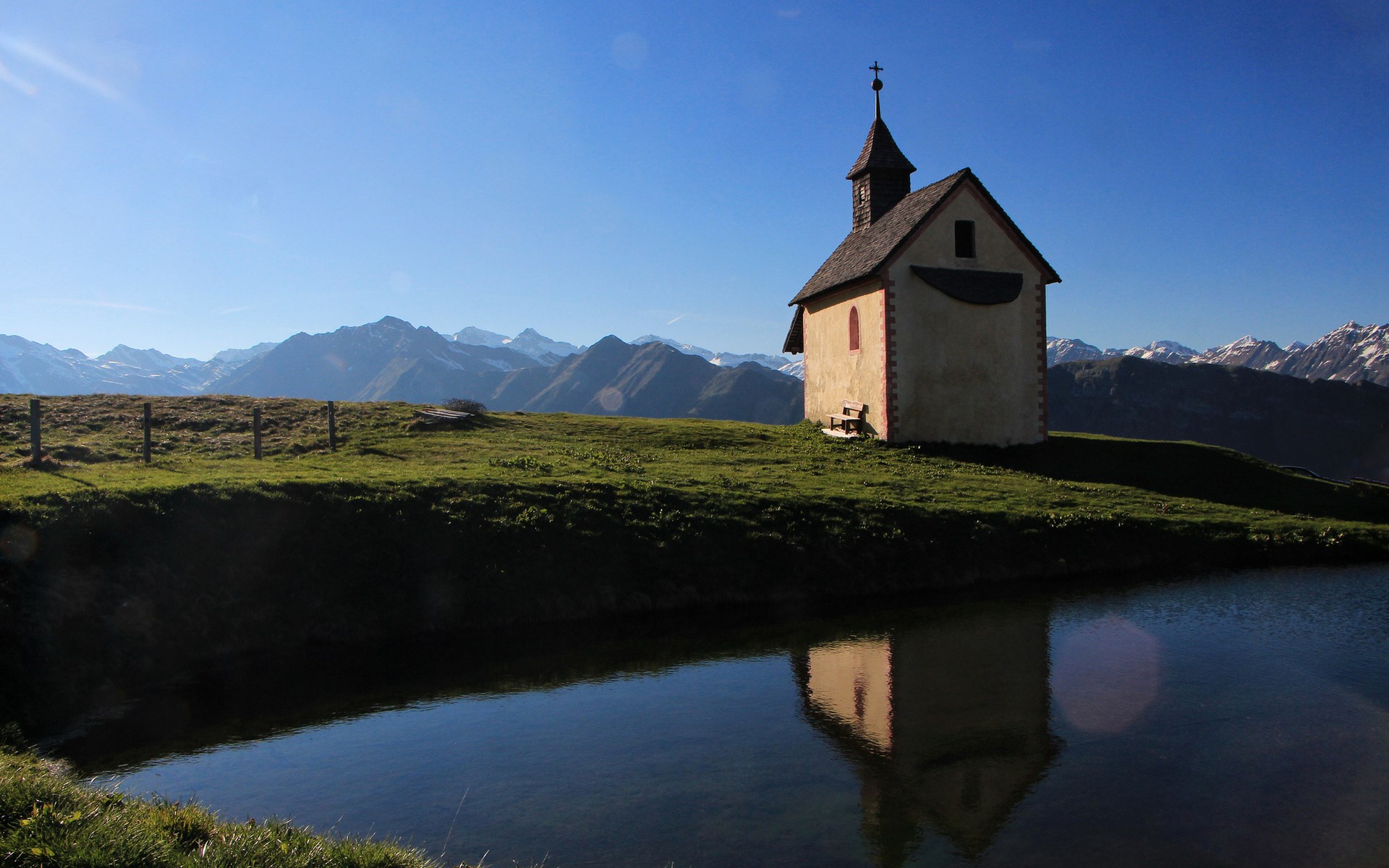 Chapel on a grassy hill overlooking mountains with a pond HD Wallpaper