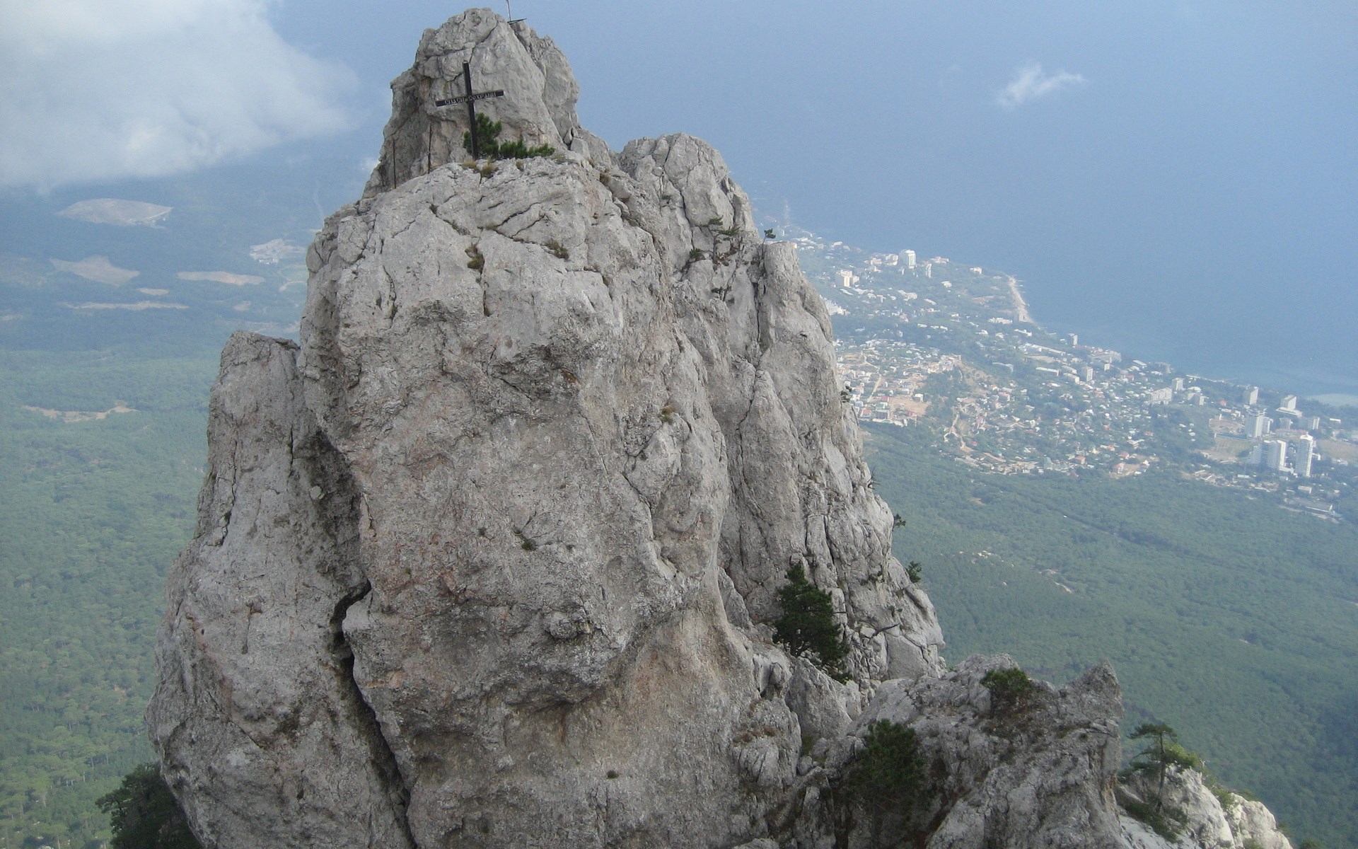 Cross on a rocky mountain peak overlooking a coastal city HD Wallpaper