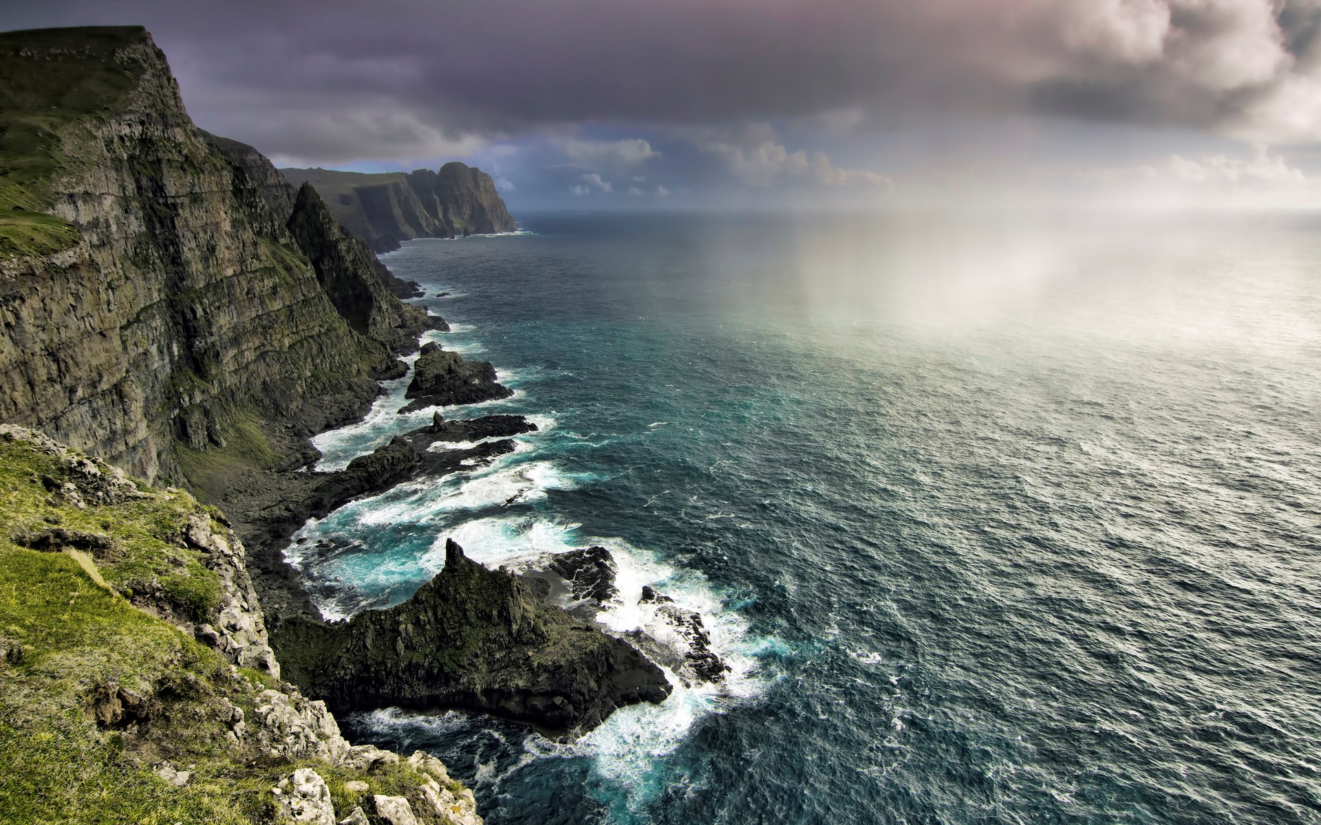 Rugged cliffs meet the turbulent ocean under a stormy sky. HD Wallpaper