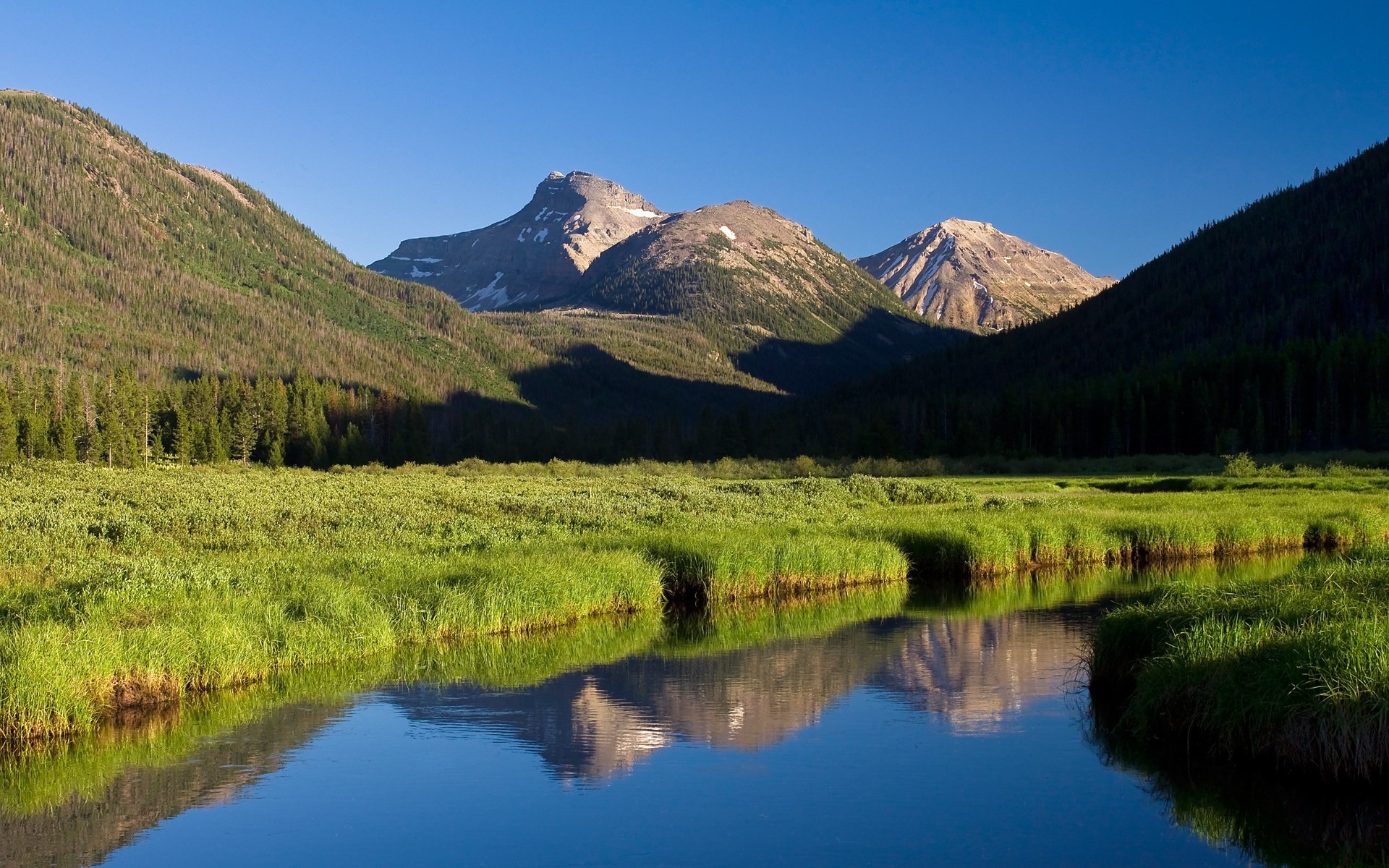 Mountain range reflected in a calm river with green grass HD Wallpaper