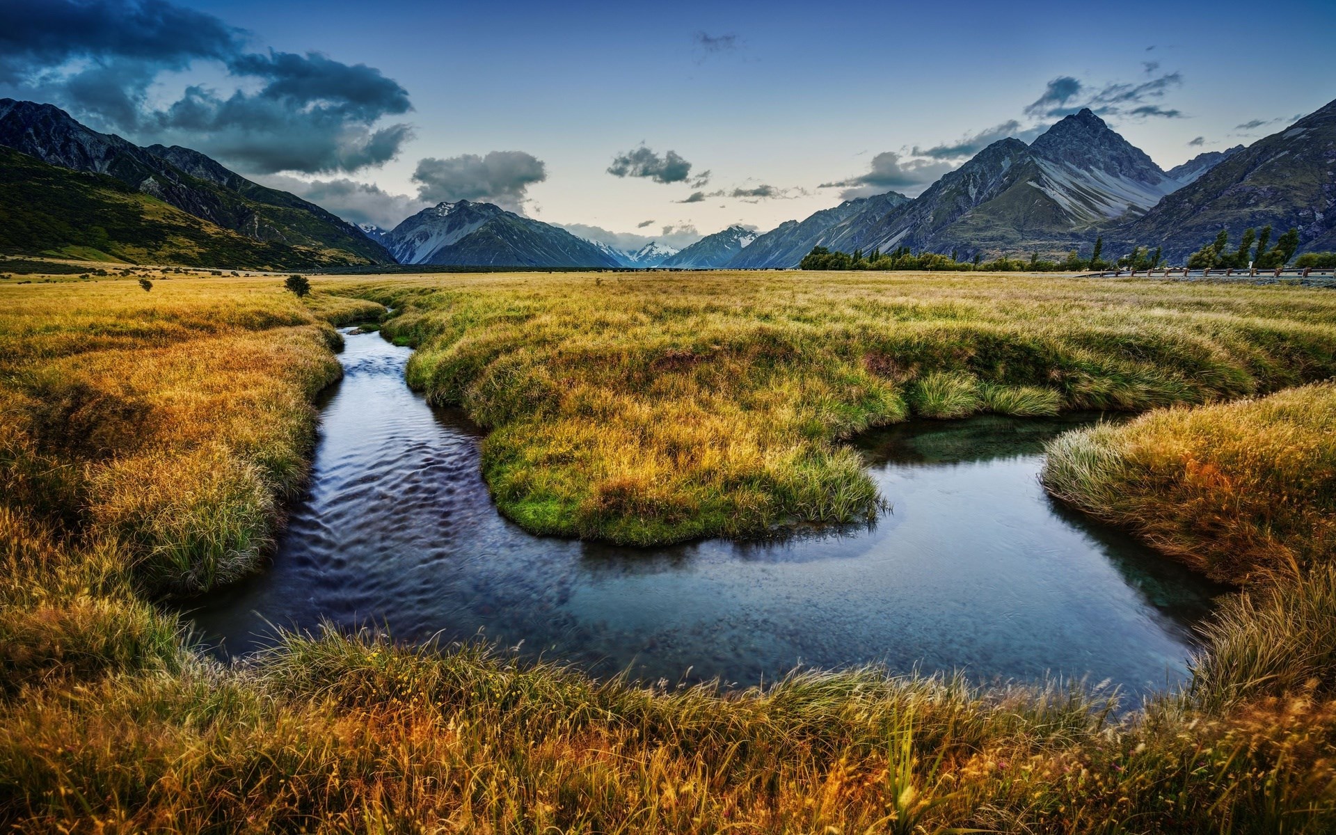 A winding stream flows through a golden meadow with mountains in the background. HD Wallpaper