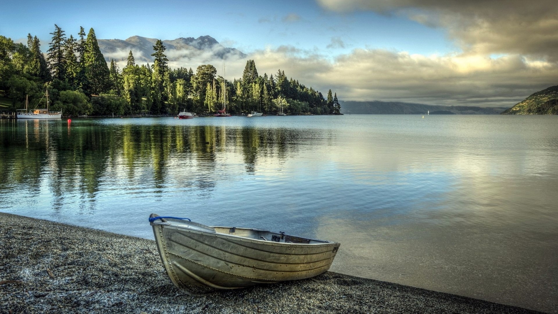 Boat on a pebble beach by a lake with trees and mountains HD Wallpaper
