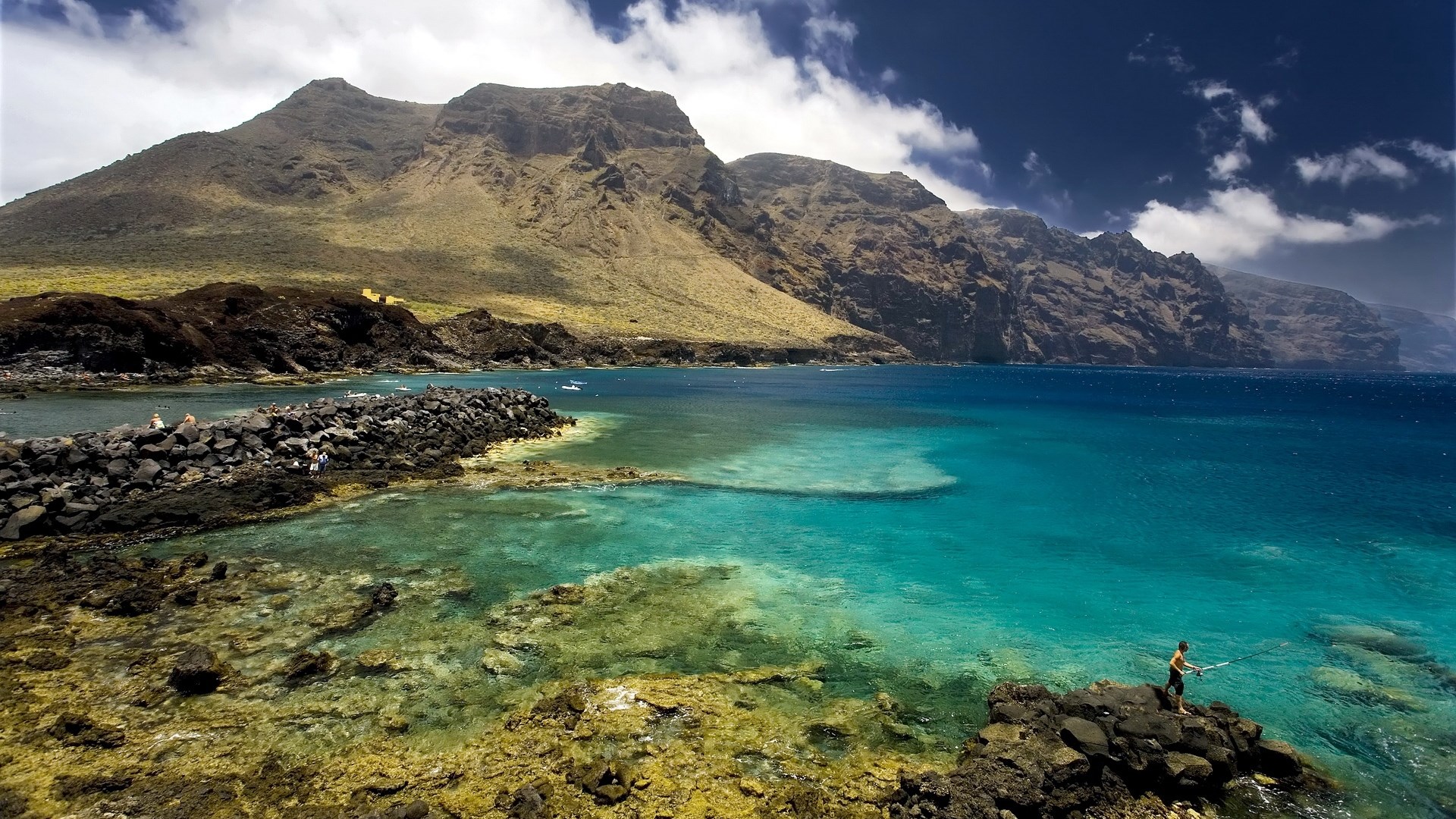 Man fishing on rocky coast with mountains and ocean HD Wallpaper