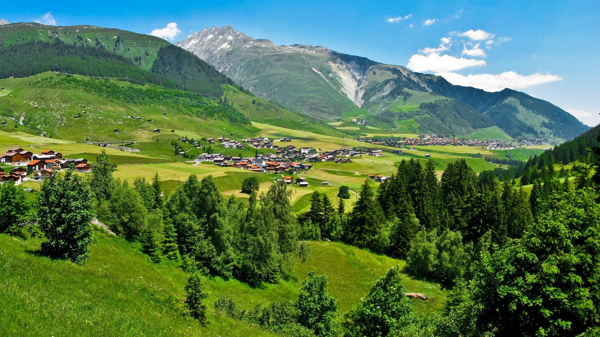 Alpine village nestled in a green valley with mountains HD Wallpaper