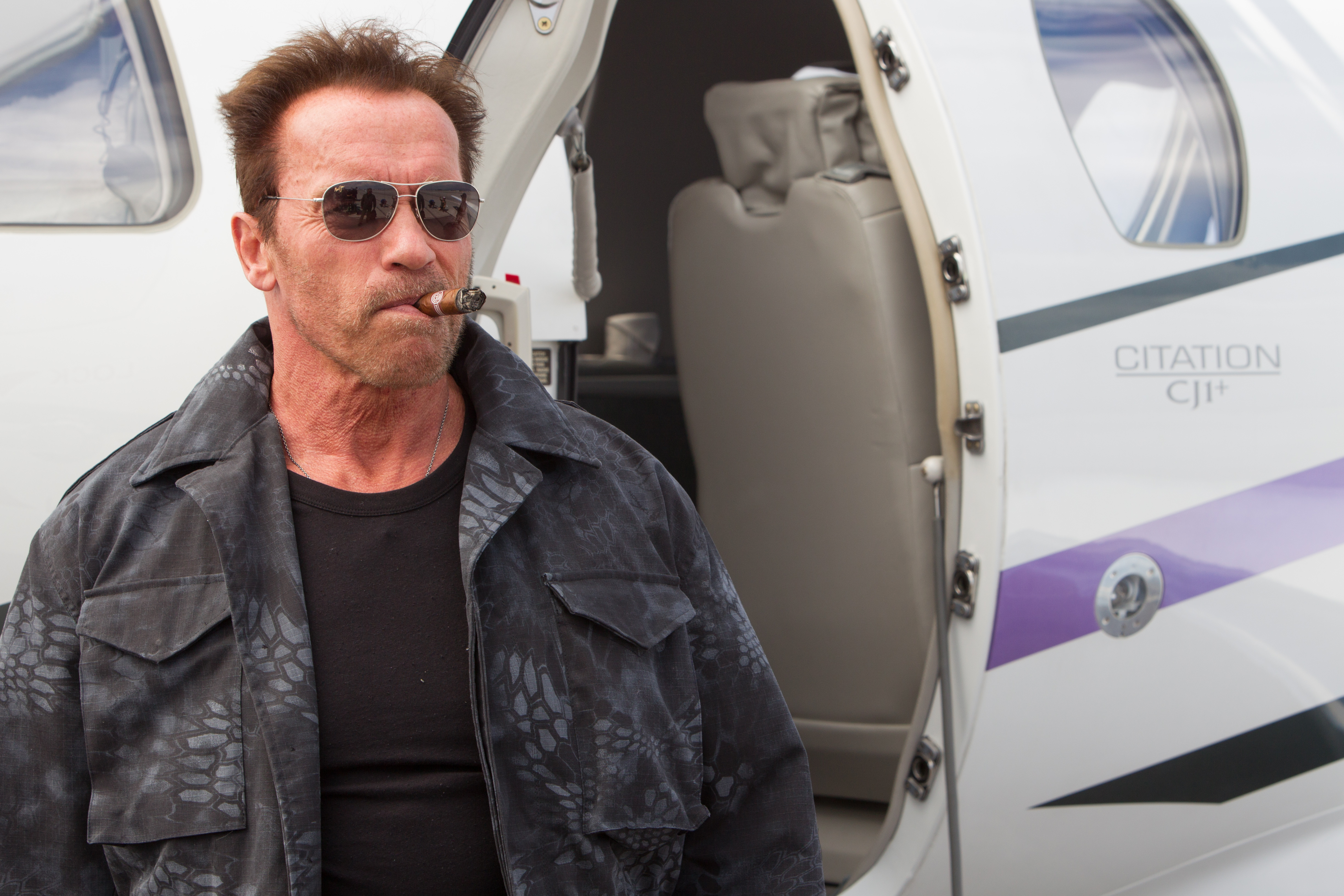 Arnold Schwarzenegger smoking a cigar by a private jet HD Wallpaper