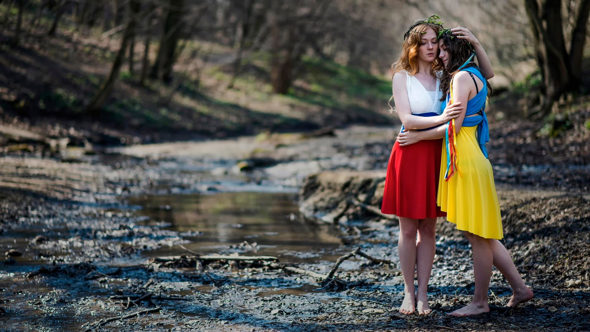 Two women embracing in a forest by a stream HD Wallpaper