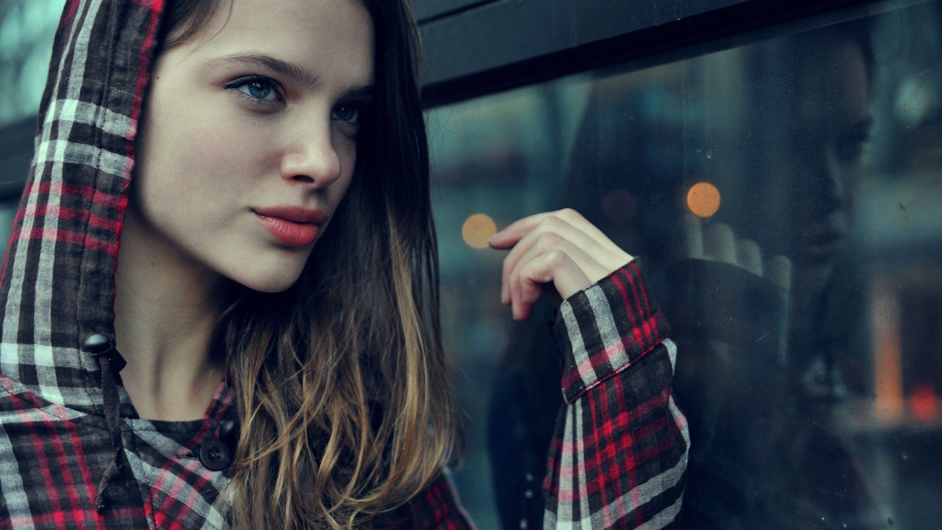 Young woman in a plaid hoodie looking out a window HD Wallpaper