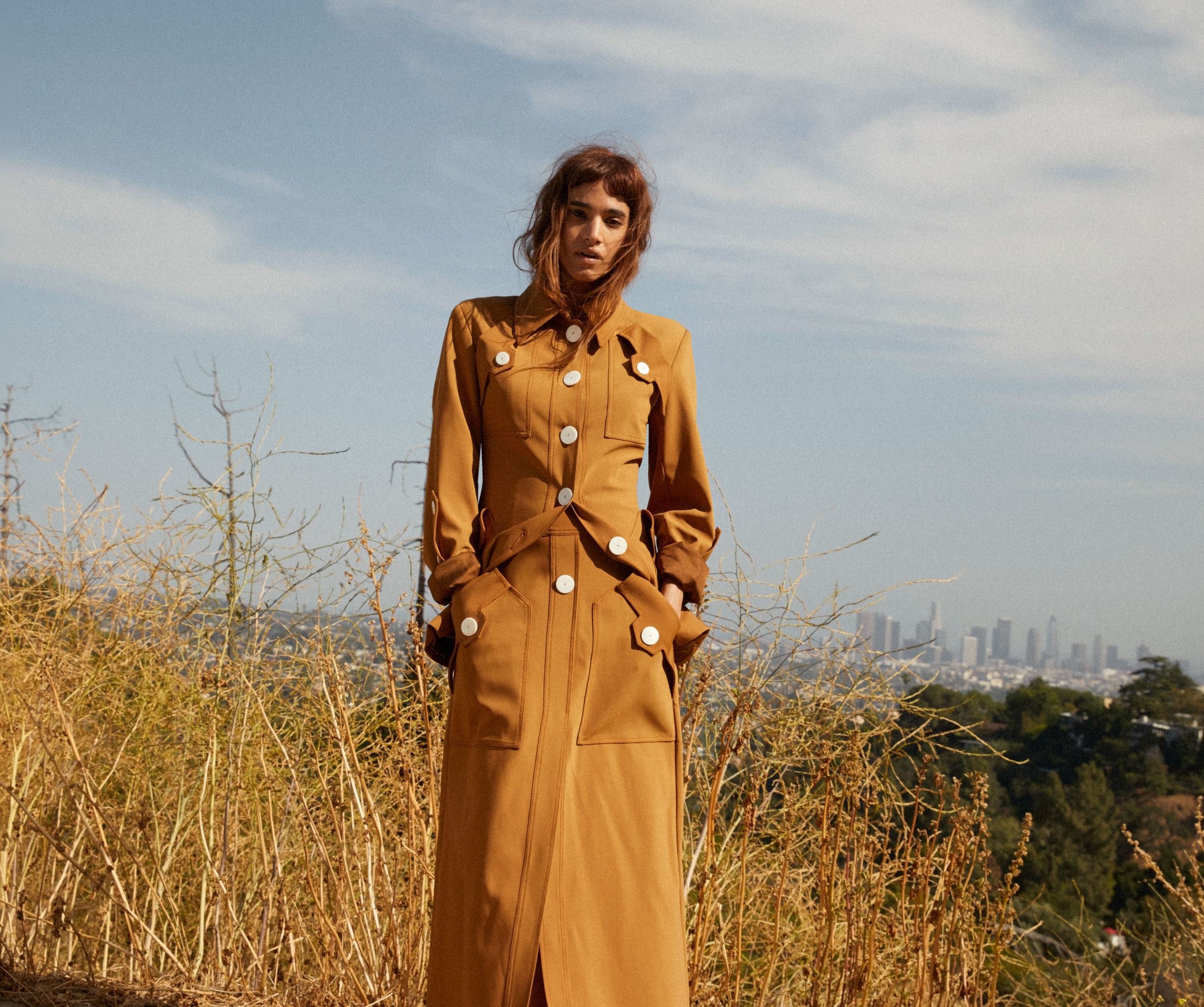 Sofia Boutella in a tan trench coat with a city skyline HD Wallpaper