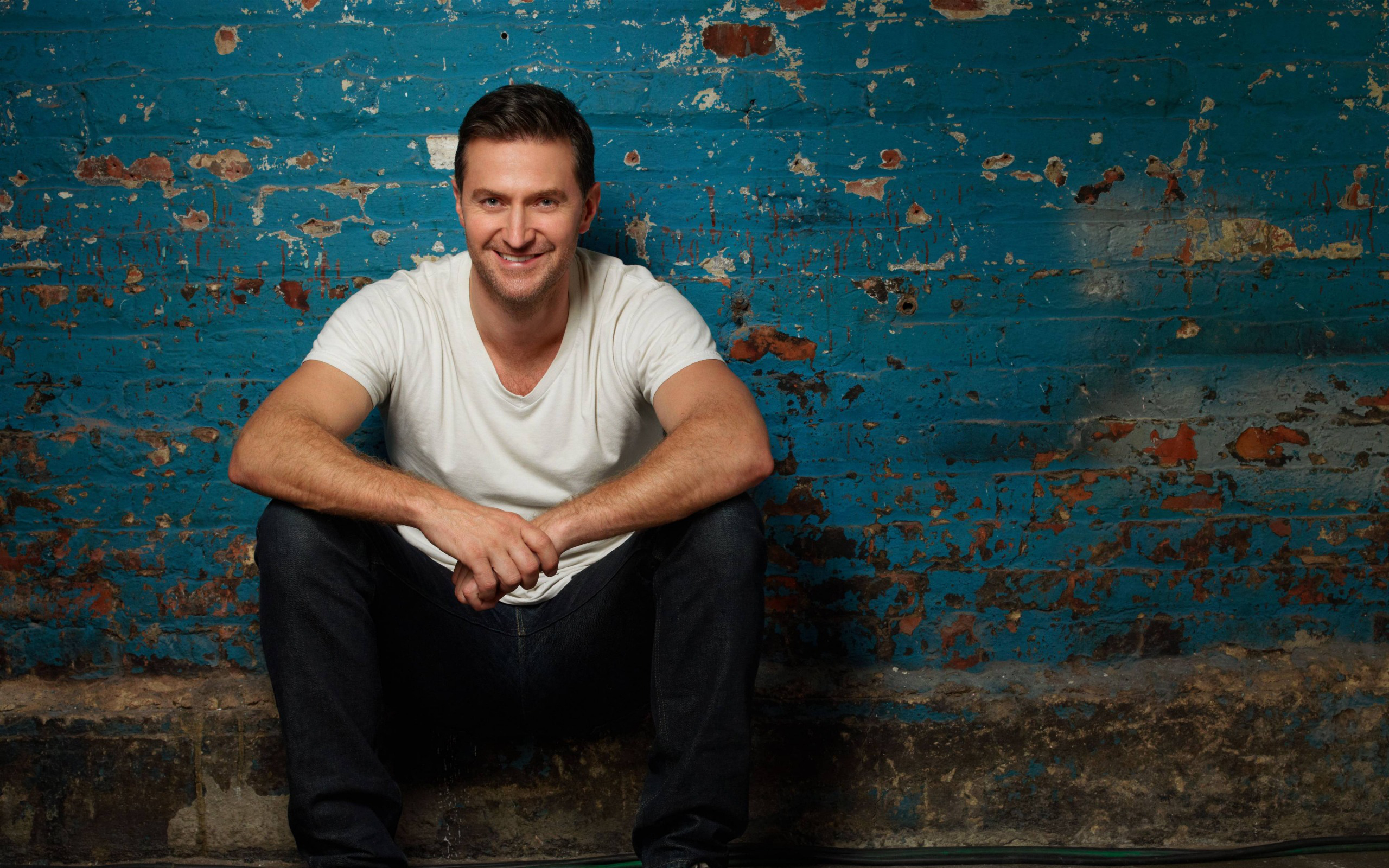 Richard Armitage sitting in front of a blue brick wall HD Wallpaper