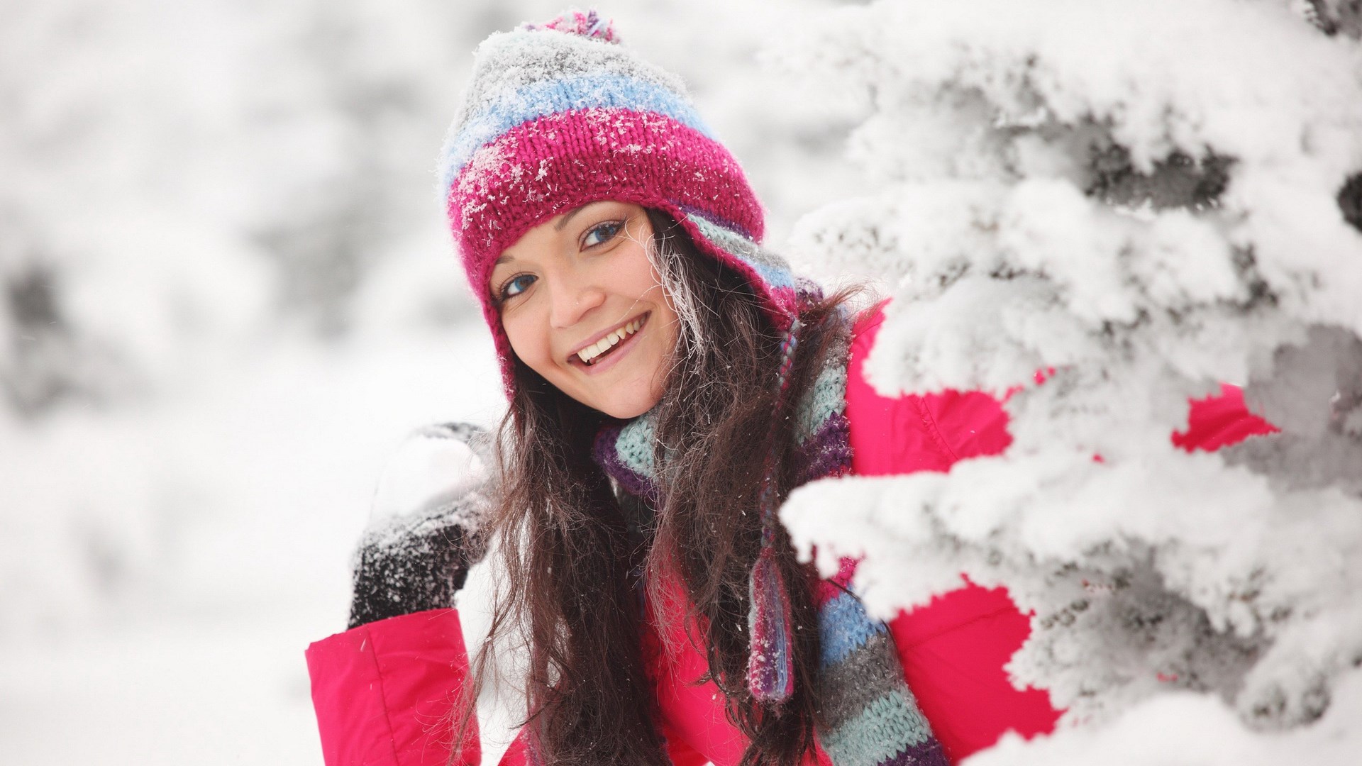 Young woman in winter clothing peeking from behind a snow-covered tree HD Wallpaper