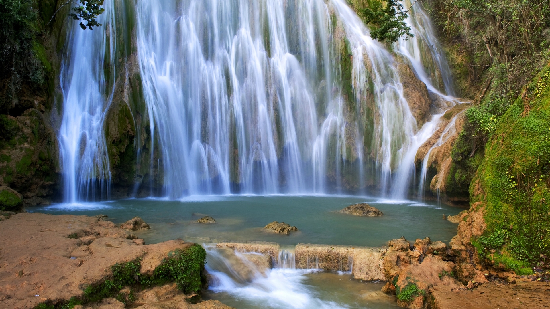 A beautiful waterfall cascading into a turquoise pool surrounded by mossy rocks and greenery. HD Wallpaper