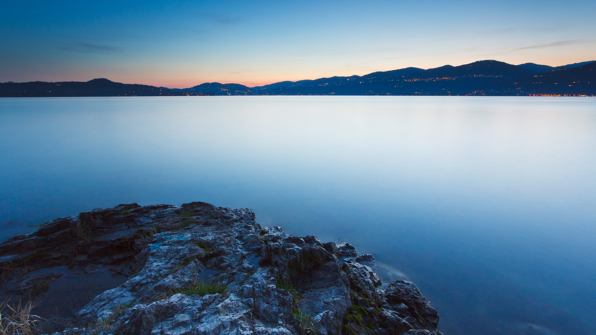 Rocky shore at dusk overlooking a calm lake and distant mountains with city lights HD Wallpaper