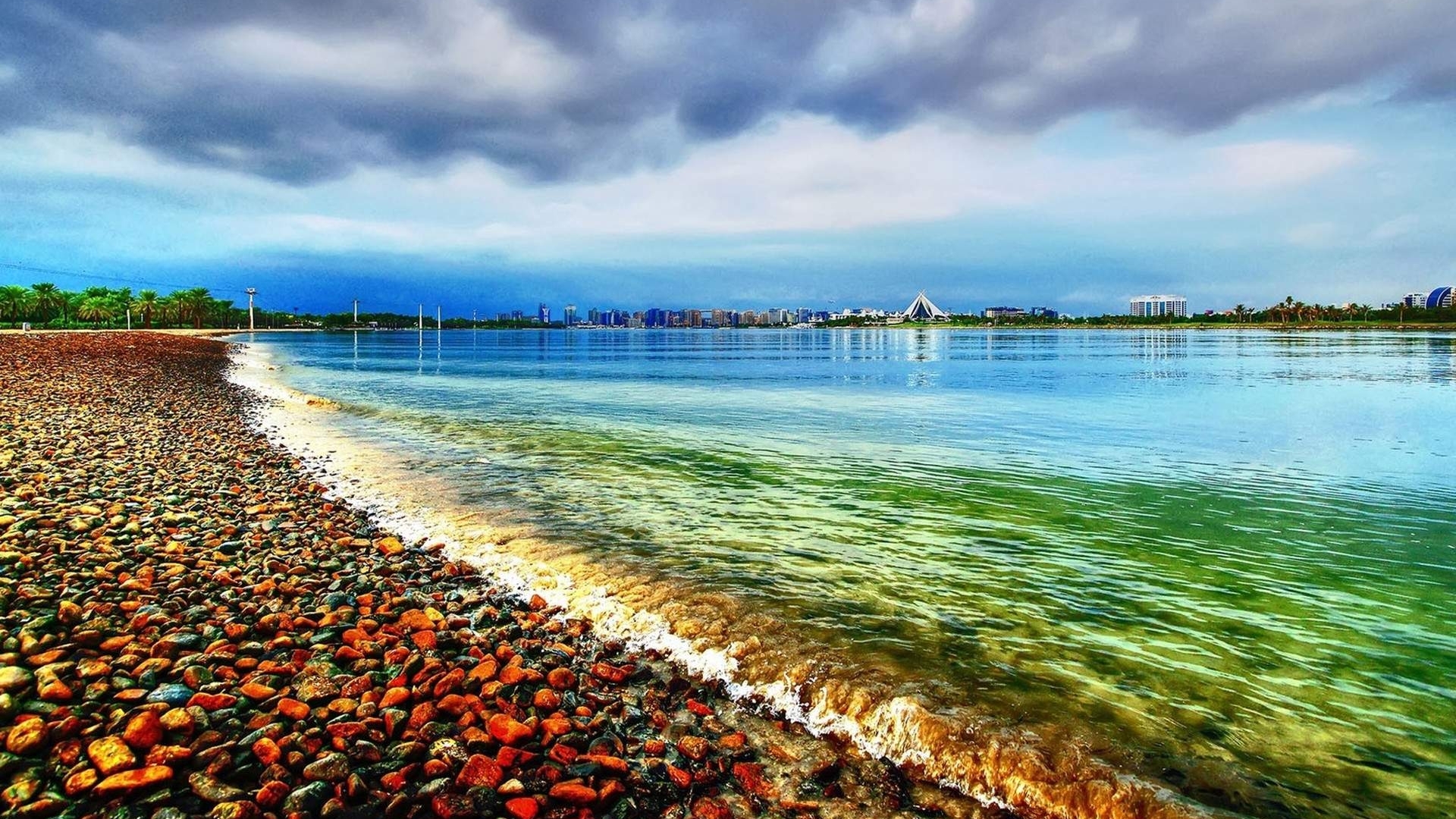 Pebble beach with calm water and city skyline HD Wallpaper