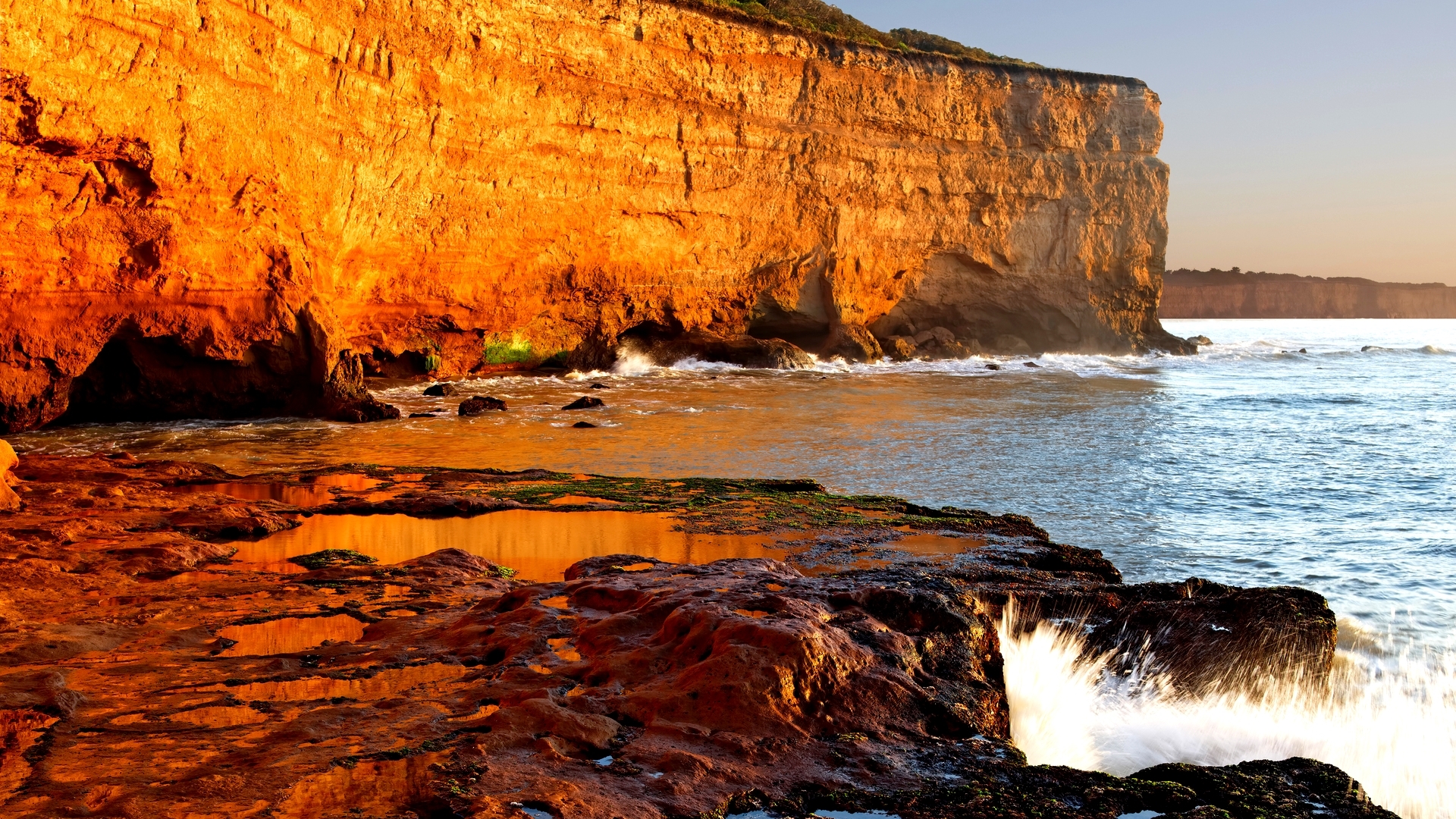 Ocean waves crashing against a rocky cliff at sunset HD Wallpaper
