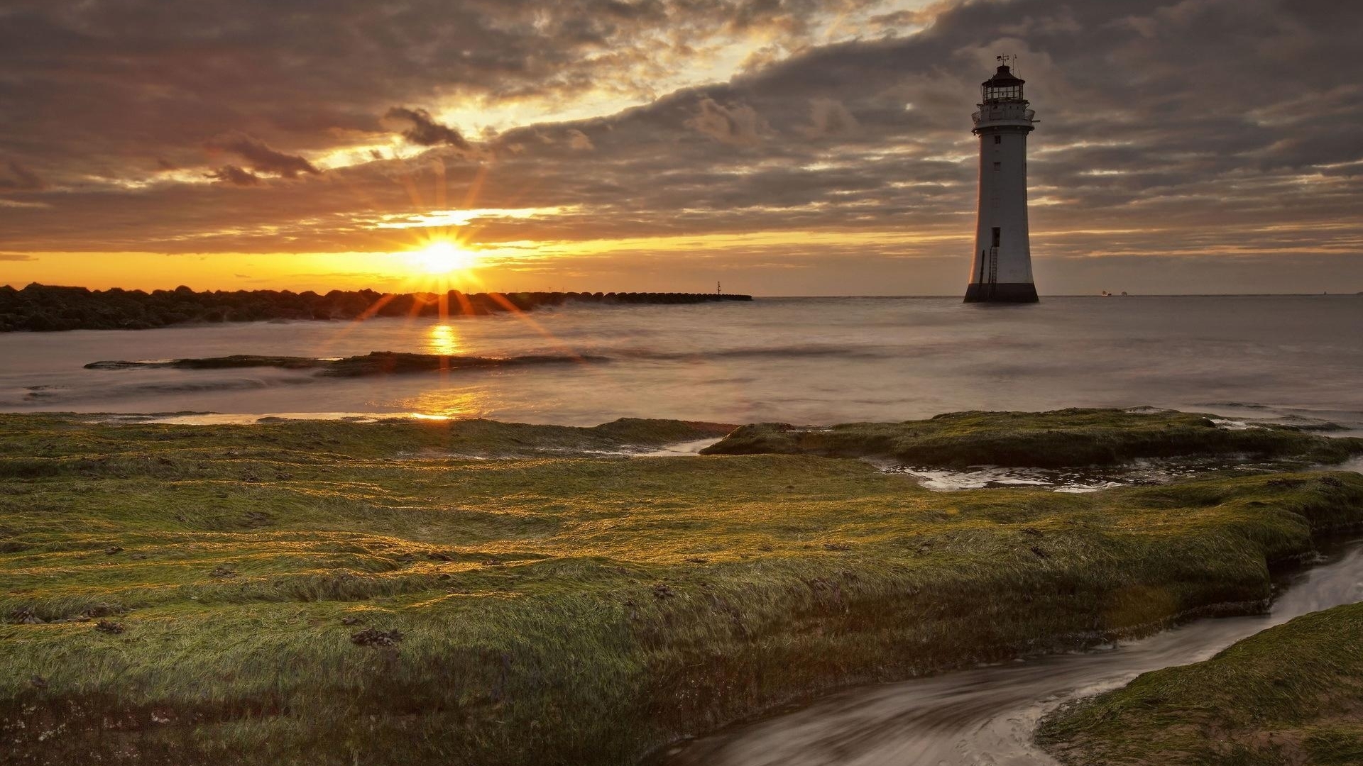 Sunset over the sea with a lighthouse and rocky shore HD Wallpaper