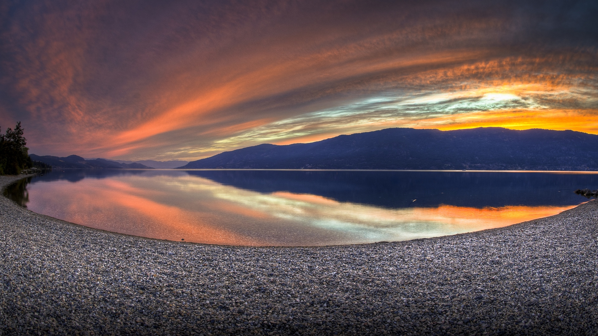 Sunset over a calm lake with mountains and a pebble beach HD Wallpaper
