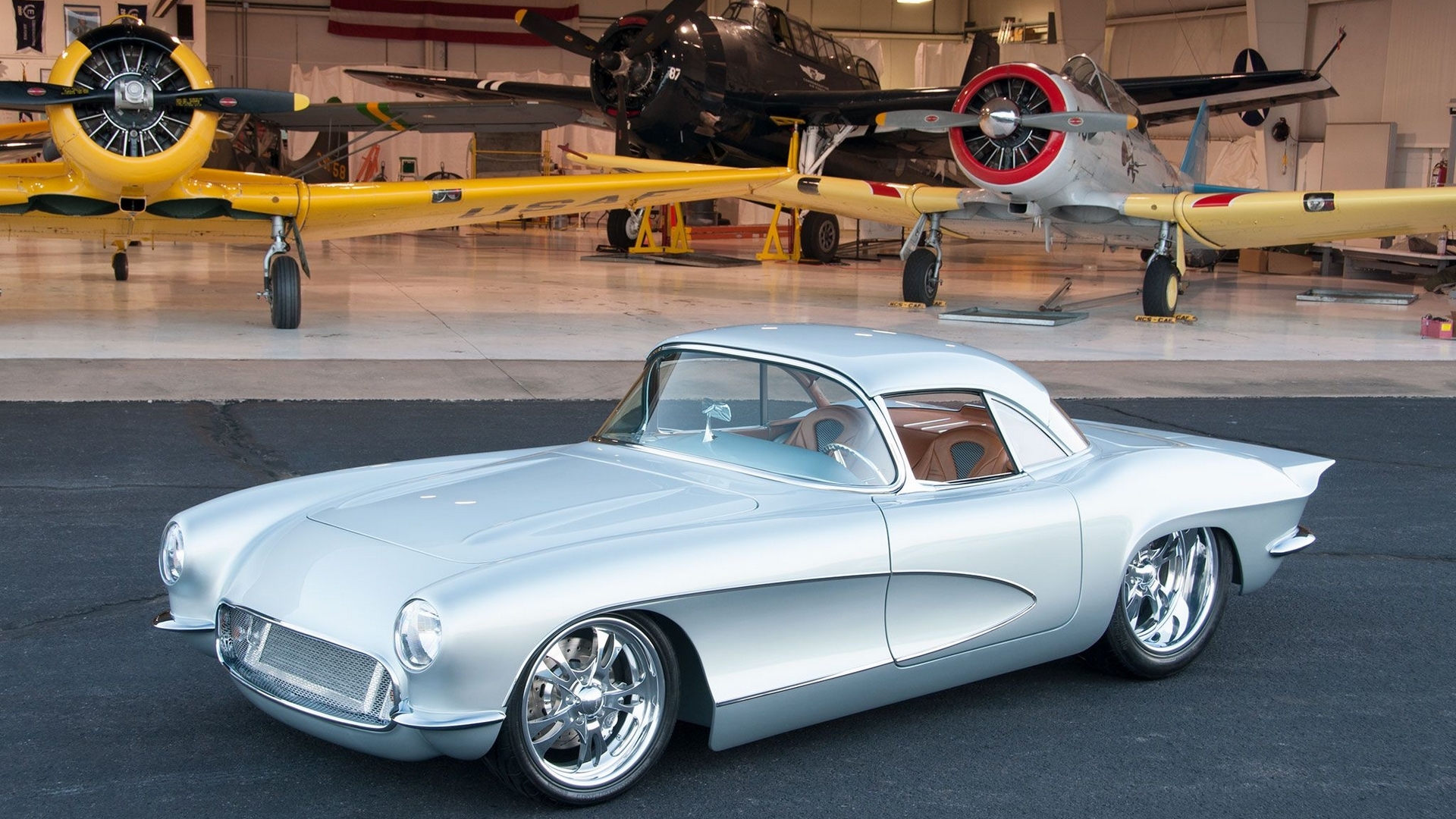 Silver 1950s Chevrolet Corvette in front of vintage airplanes HD Wallpaper