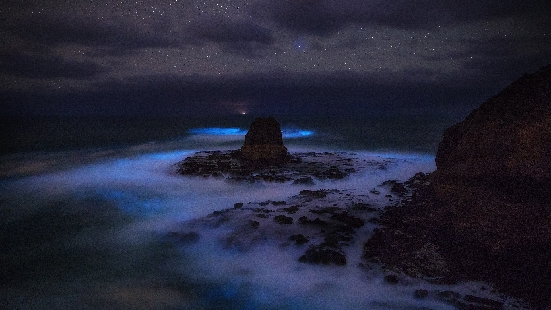 Bioluminescent ocean waves crash on rocky shore under starry night sky HD Wallpaper