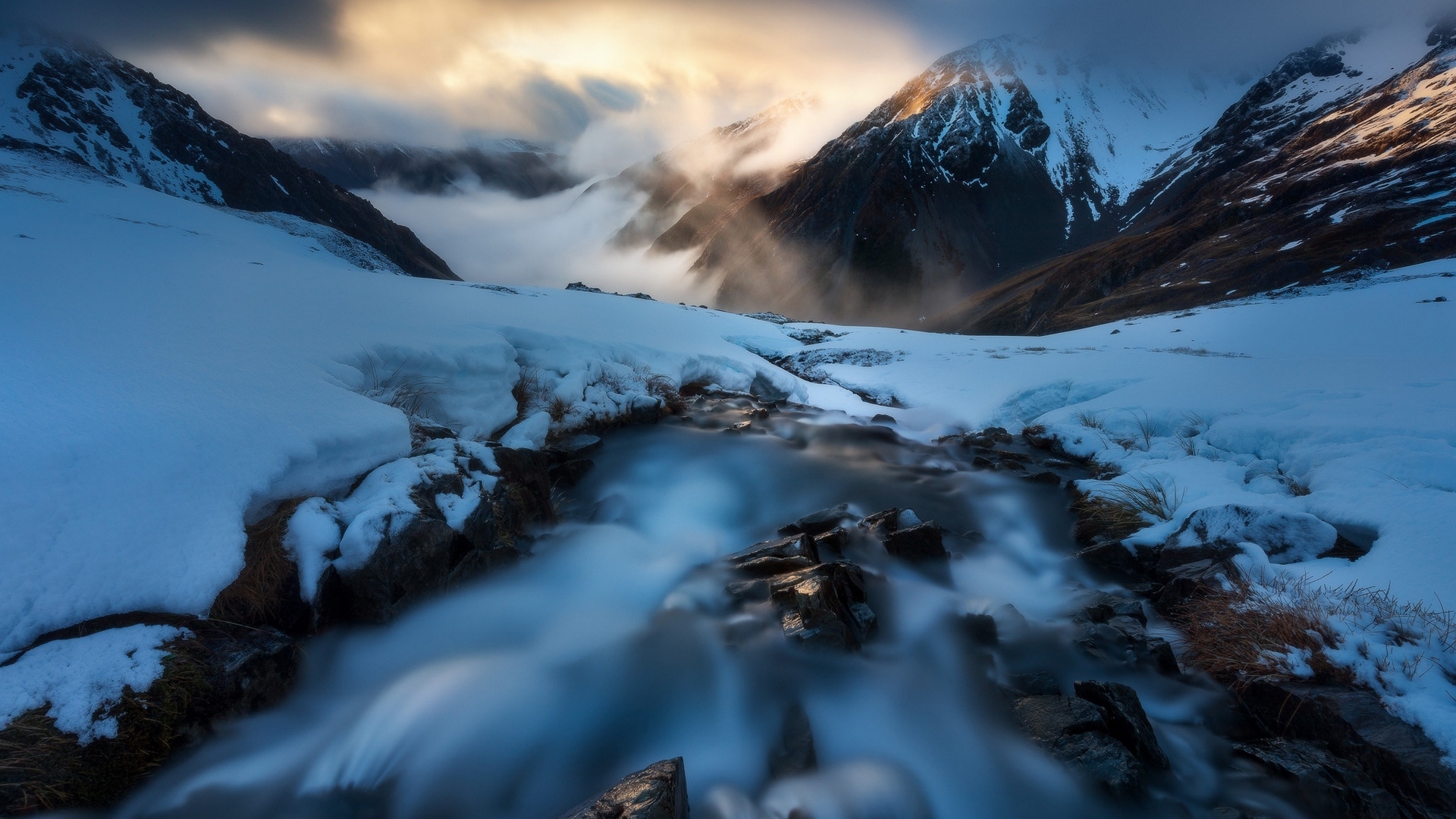 Snowy mountains with a stream and dramatic clouds HD Wallpaper