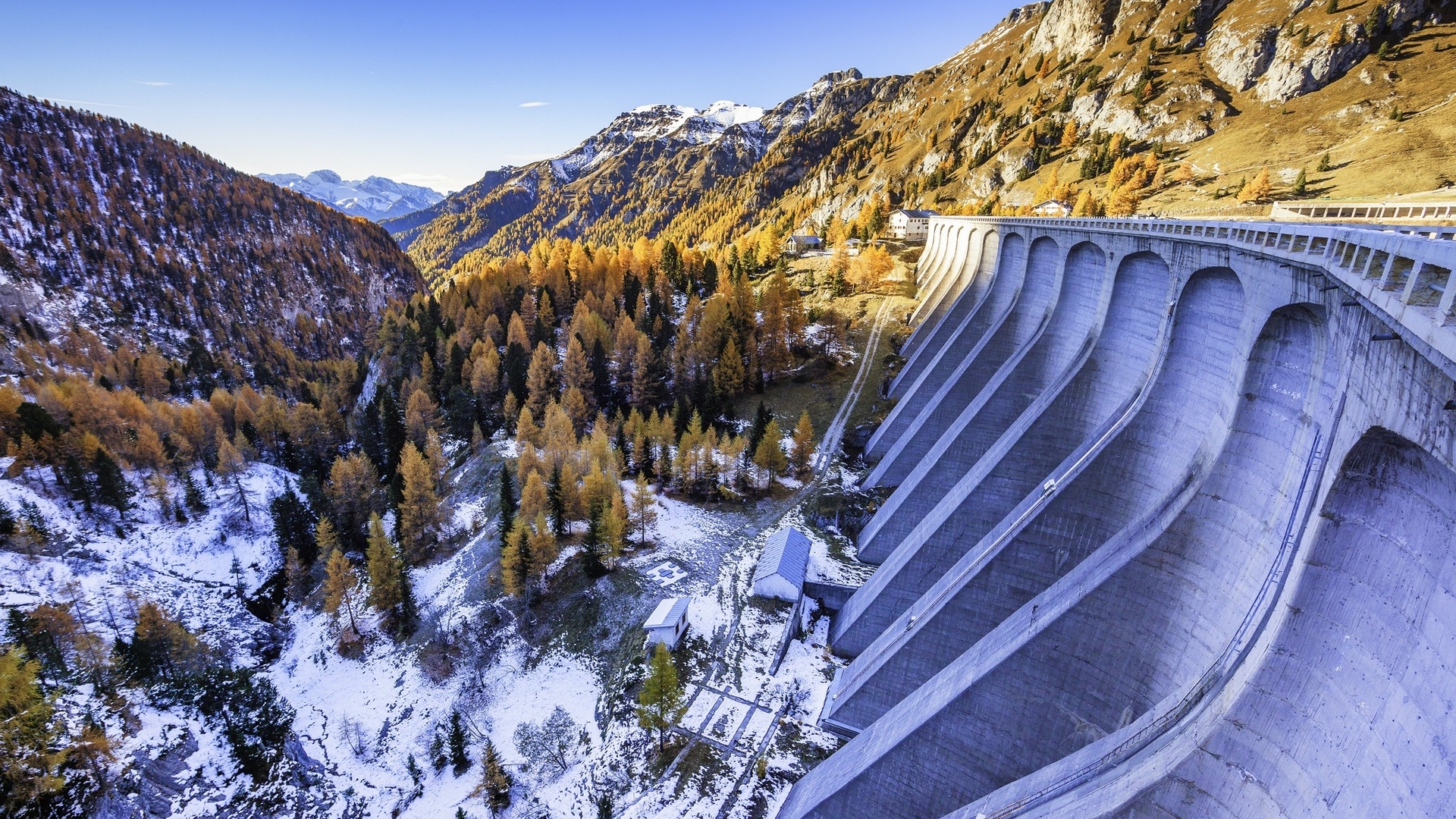 Curved concrete dam in a mountain valley with autumn trees and snow HD Wallpaper
