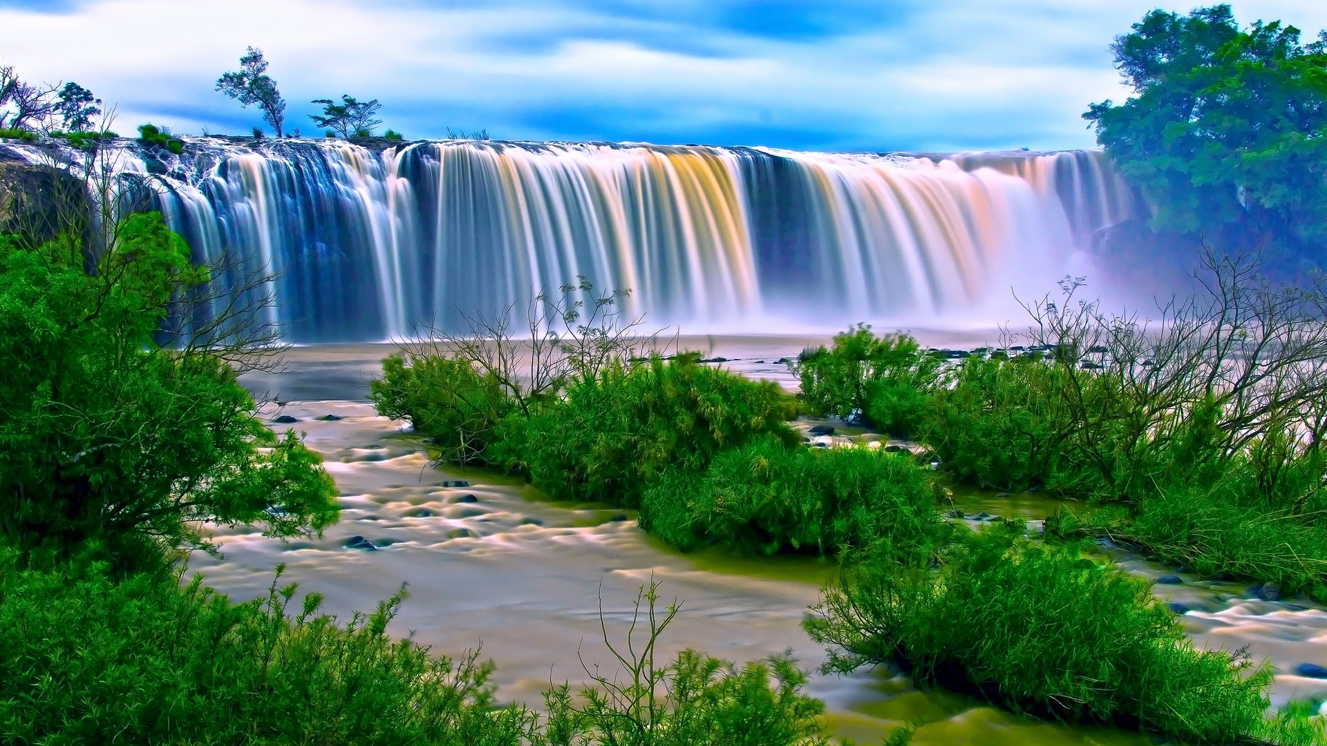 Wide waterfall cascading over a rocky ledge with green foliage HD Wallpaper
