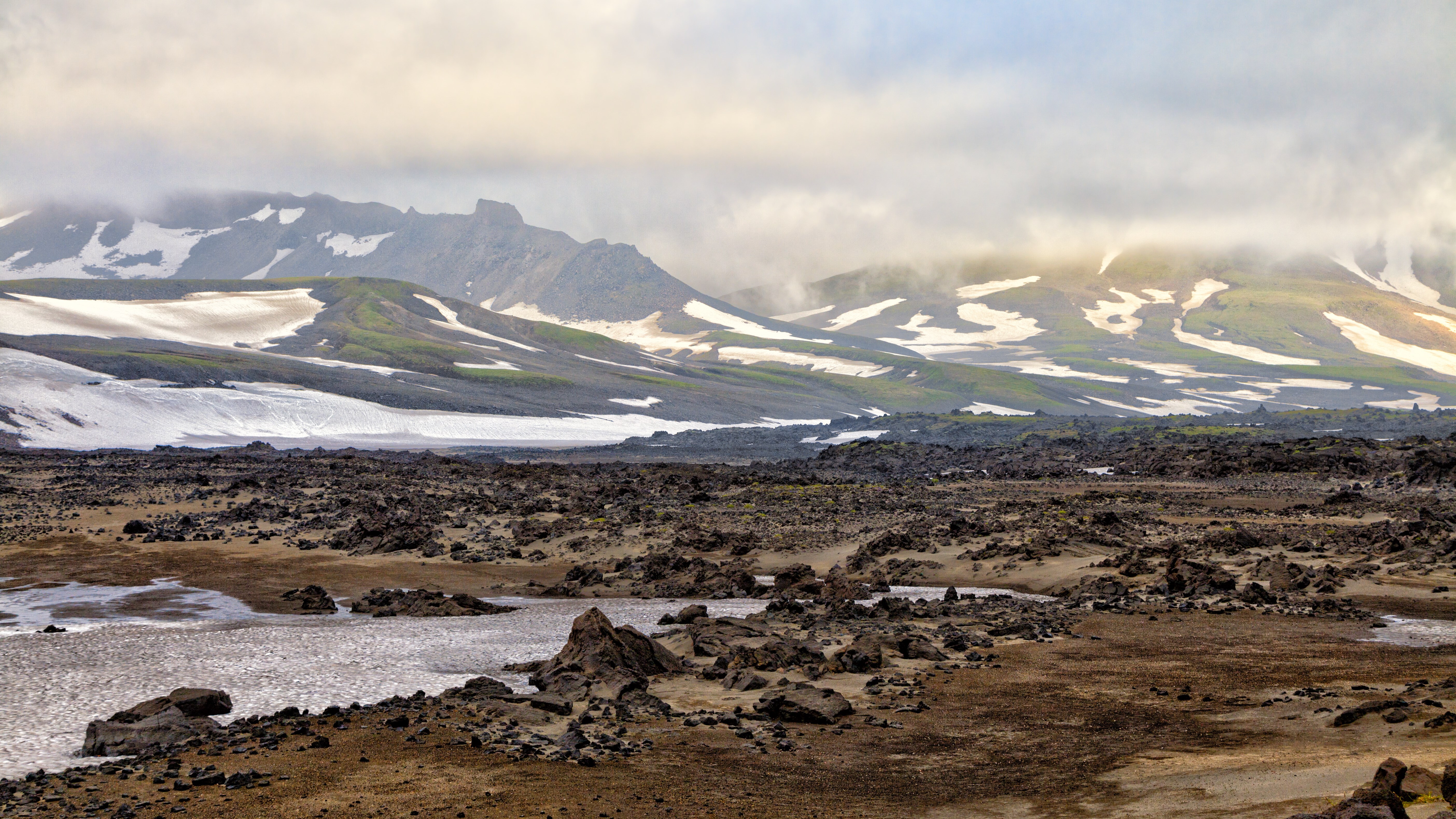 Volcanic landscape with snow-capped mountains under cloudy sky HD Wallpaper