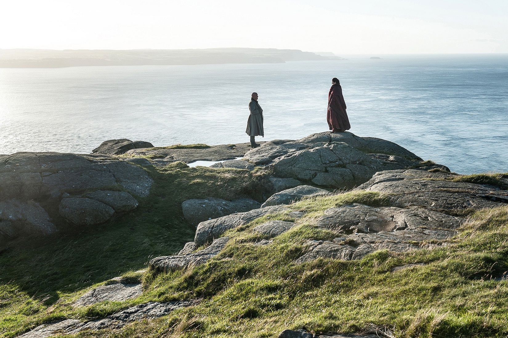 Two figures stand on a rocky coastline overlooking the sea HD Wallpaper