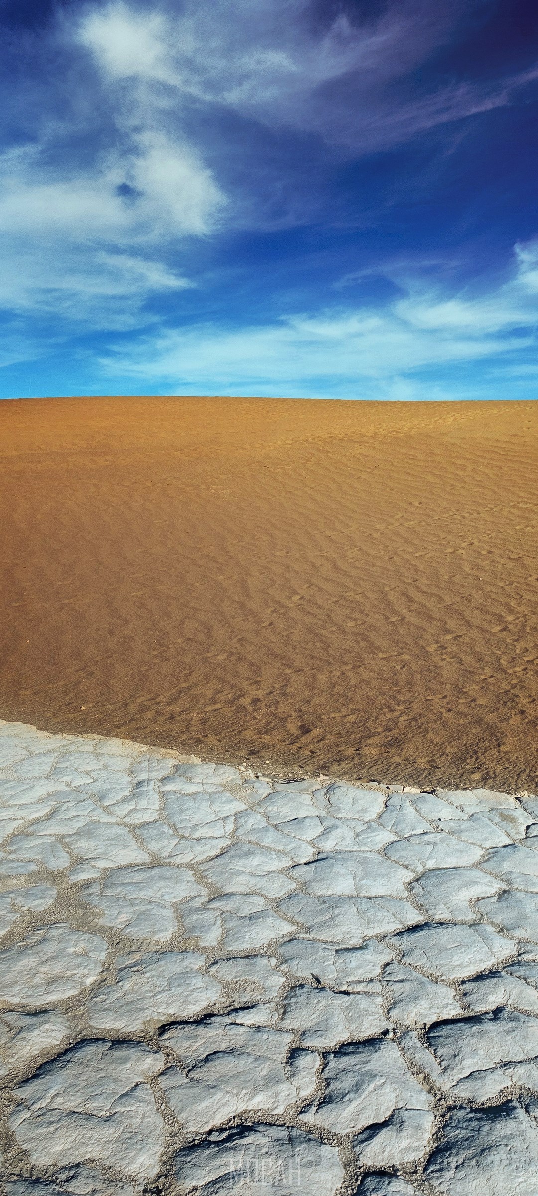 Desert landscape with cracked earth and sand dunes under a blue sky HD Phone Wallpaper