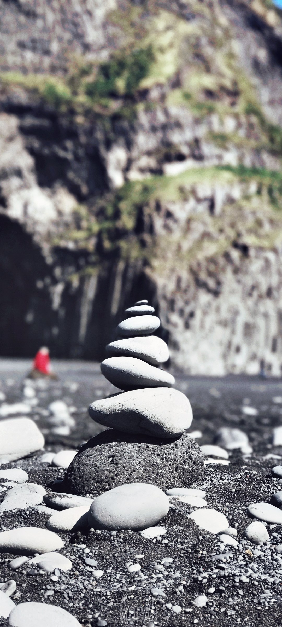 Stacked stones on a black sand beach with a person in red HD Phone Wallpaper