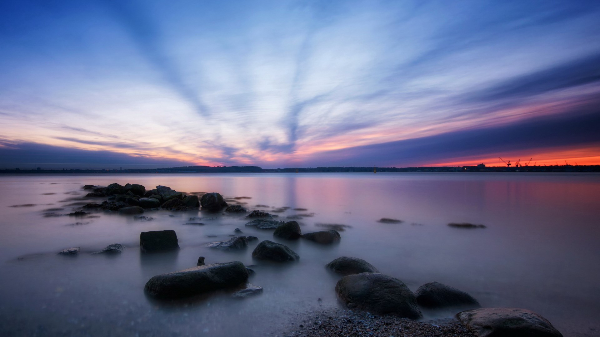 Sunset over a rocky shore with cranes in the distance HD Wallpaper