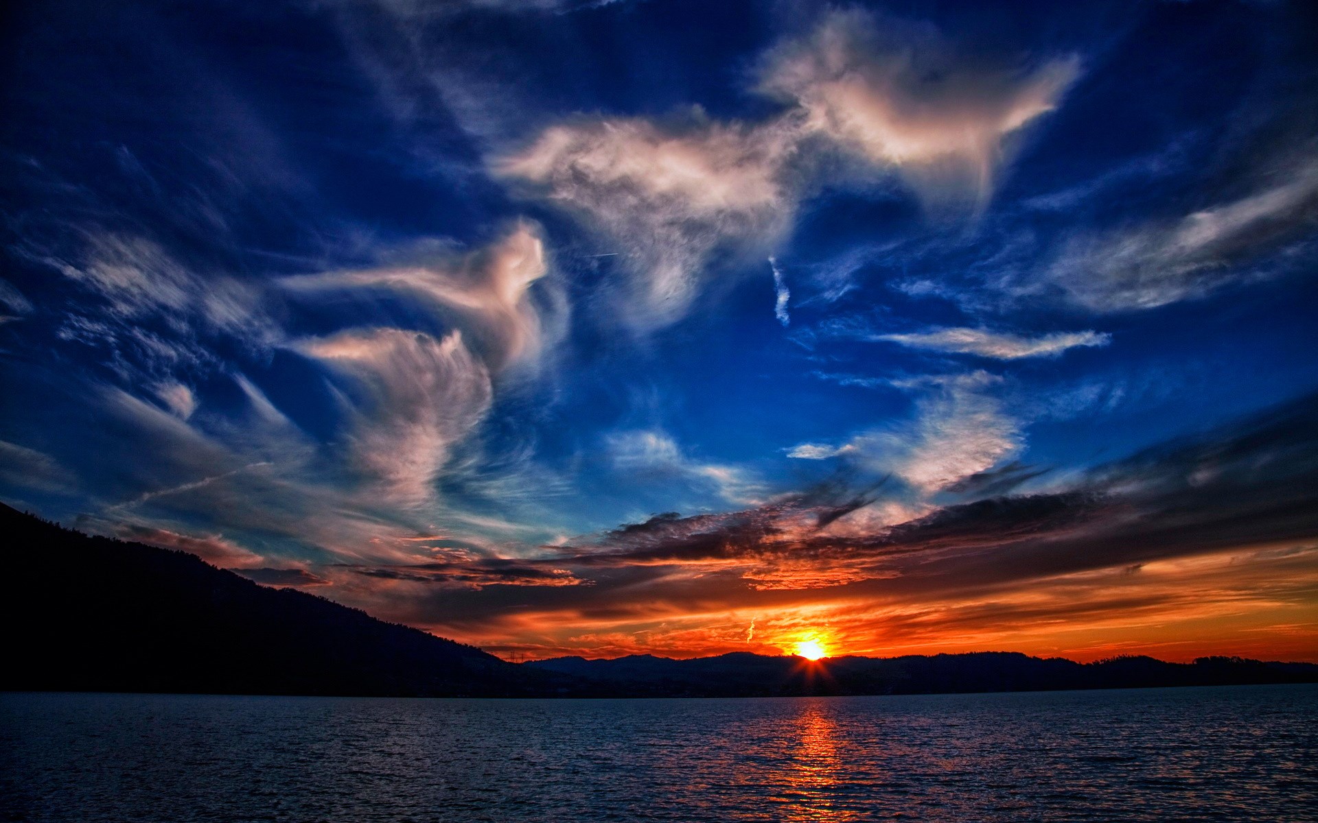 Sunset over a calm body of water with silhouetted mountains and dramatic clouds HD Wallpaper