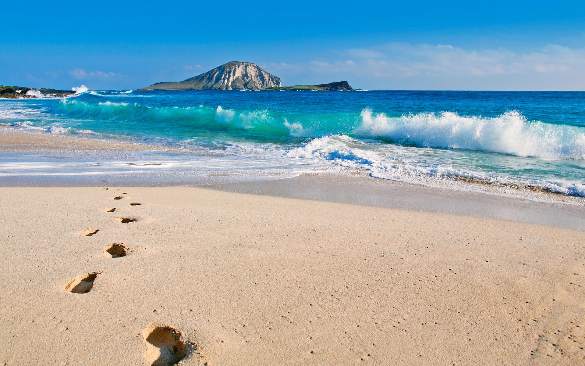 Footprints on a sandy beach with waves and an island HD Wallpaper