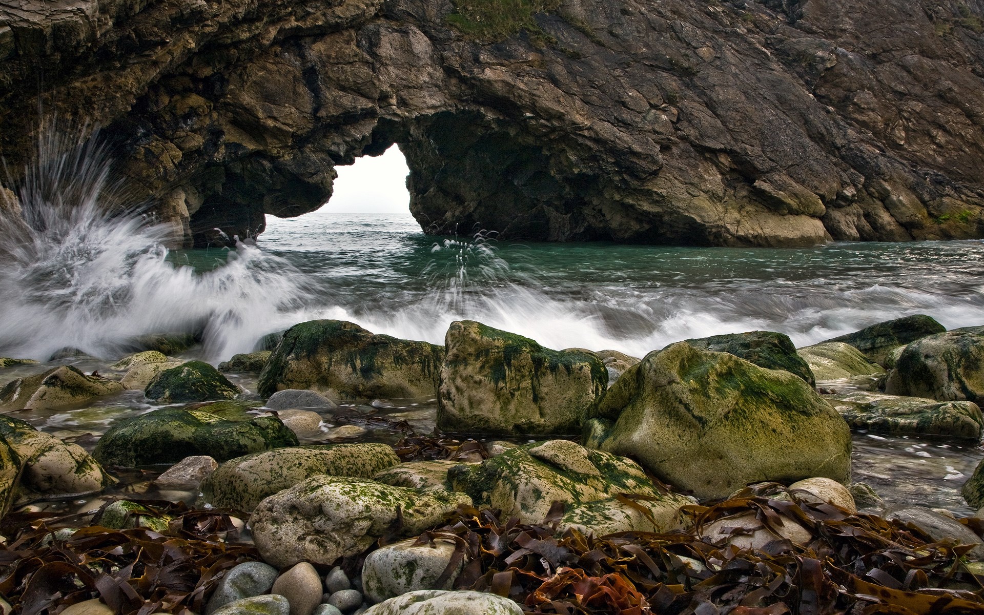 Waves crashing through a rock archway on a rocky shore HD Wallpaper
