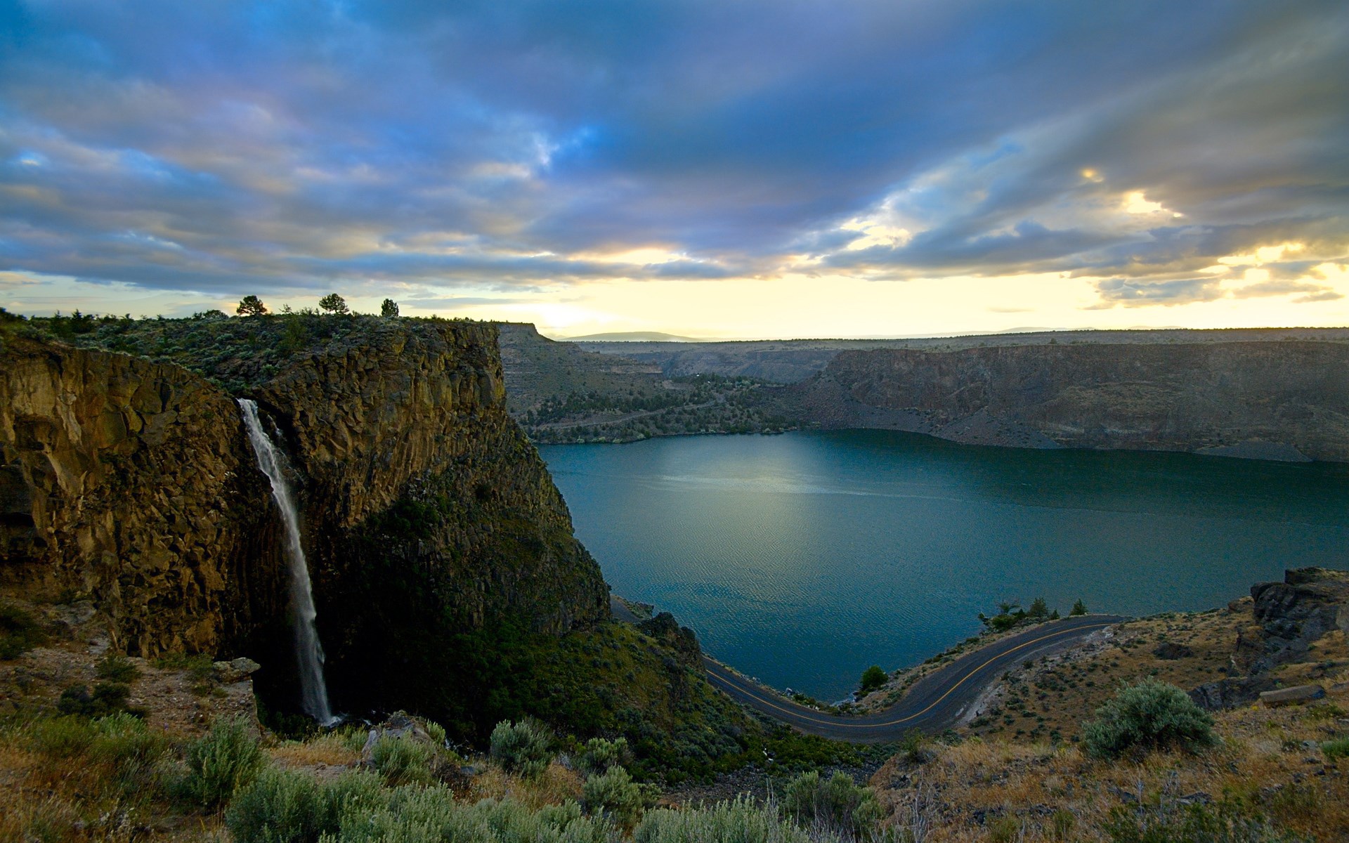 Waterfall cascading into a lake with a winding road HD Wallpaper